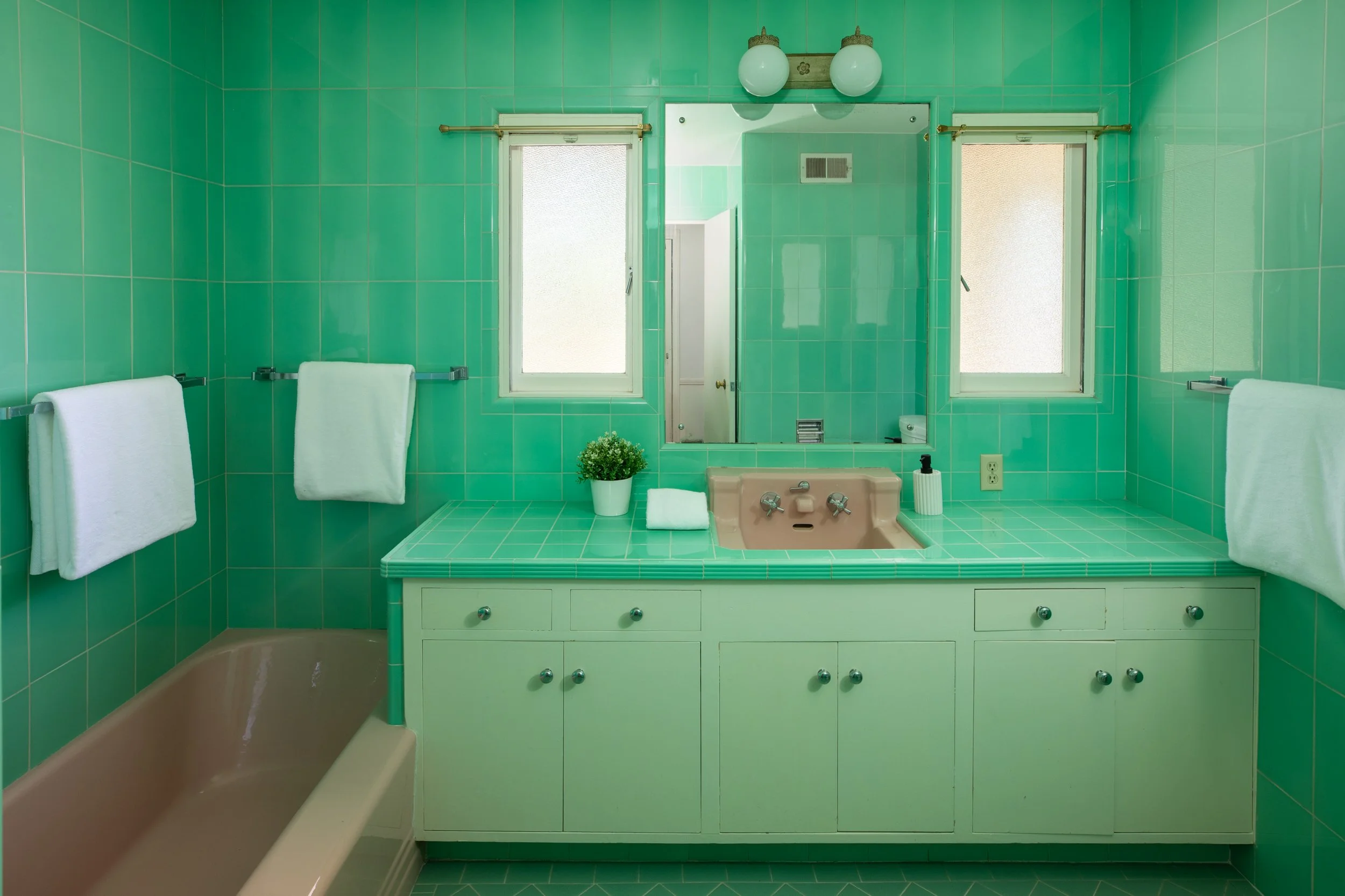 A vintage bathroom with green tiles and a pink sink, featuring a large mirror, two windows, white towels, a small potted plant, and a beige bathtub.