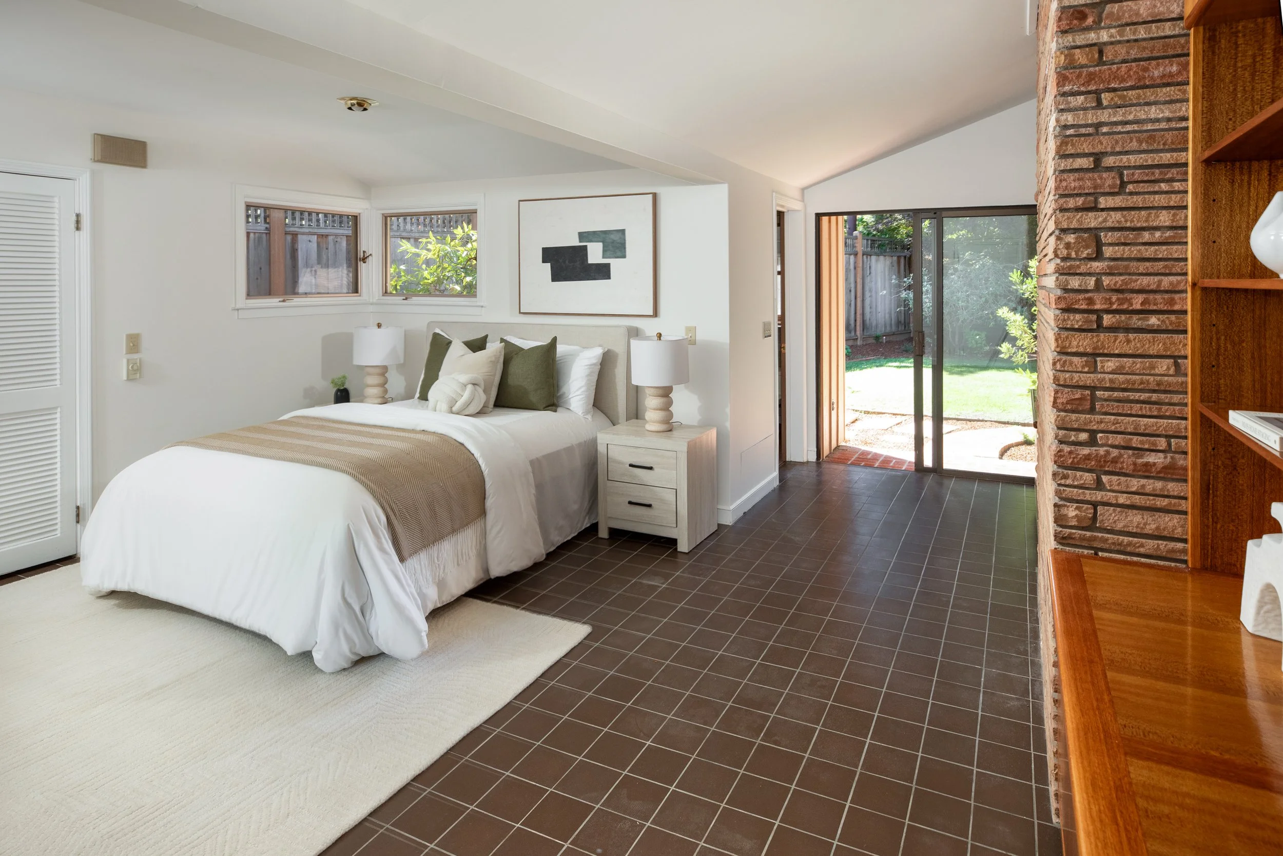 A bedroom with a bed, nightstands, lamps, and artwork, with a sliding glass door leading to a backyard.