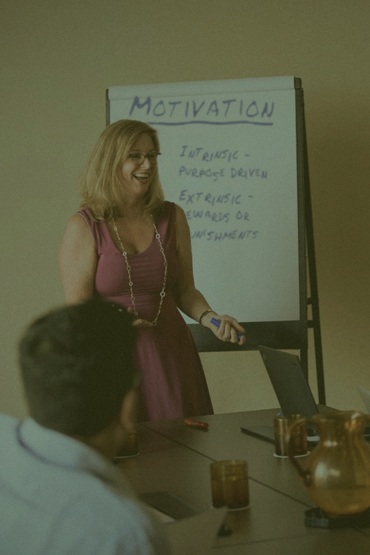 A woman standing in front of a whiteboard with the word "Motivation" written on it, giving a presentation or leading a discussion in a meeting room.