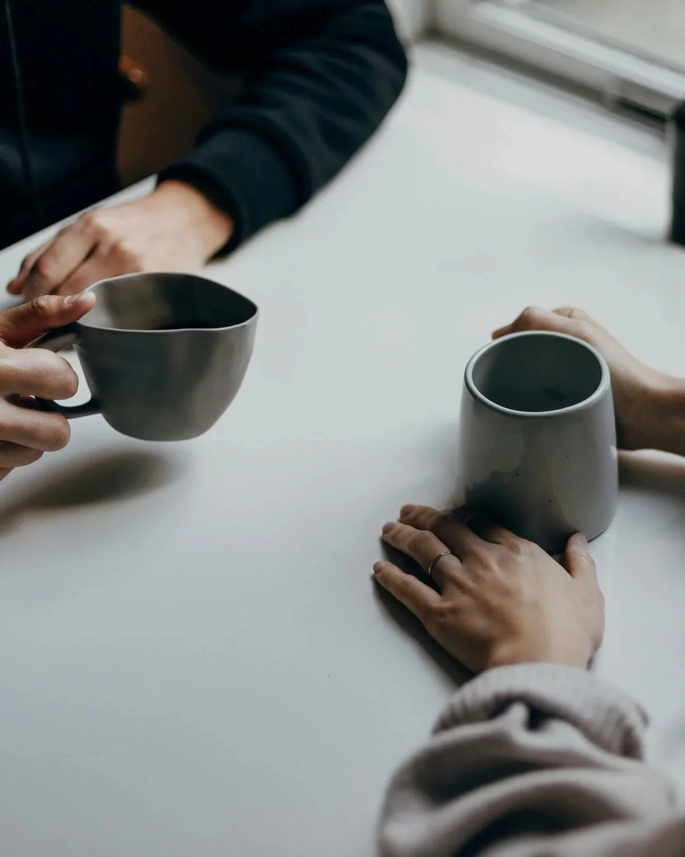 A ghostwriter and an author sitting at a white table, each holding a different mug, one black and the other gray, in a well-lit indoor space.