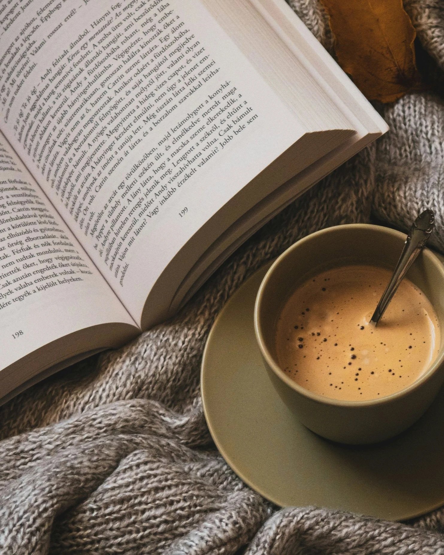 A cup of coffee with foam and foam art, placed on a saucer, alongside an open ghostwritten book resting on a cozy, chunky gray knit blanket.