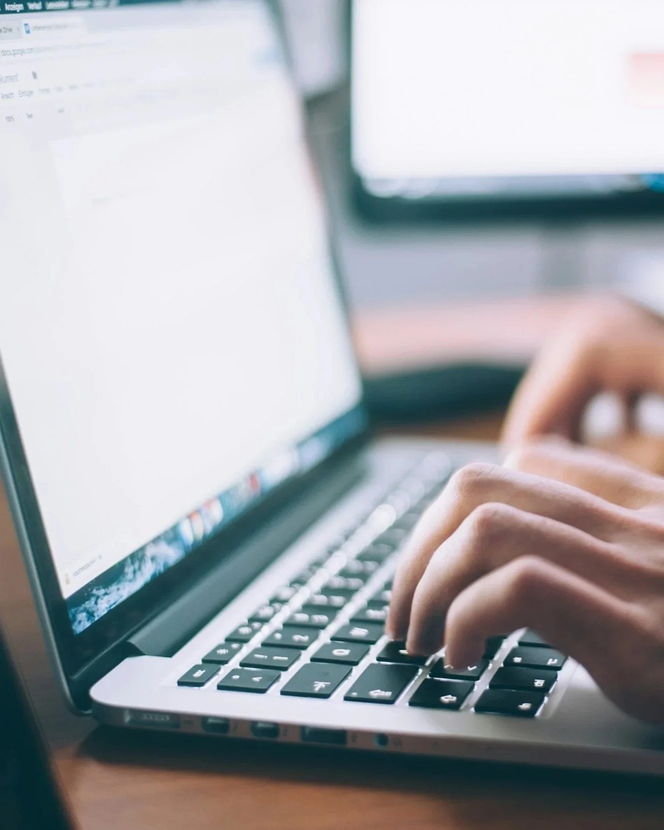 Close-up of a ghostwriters hands typing on a laptop keyboard with a blurred computer screen in the background.