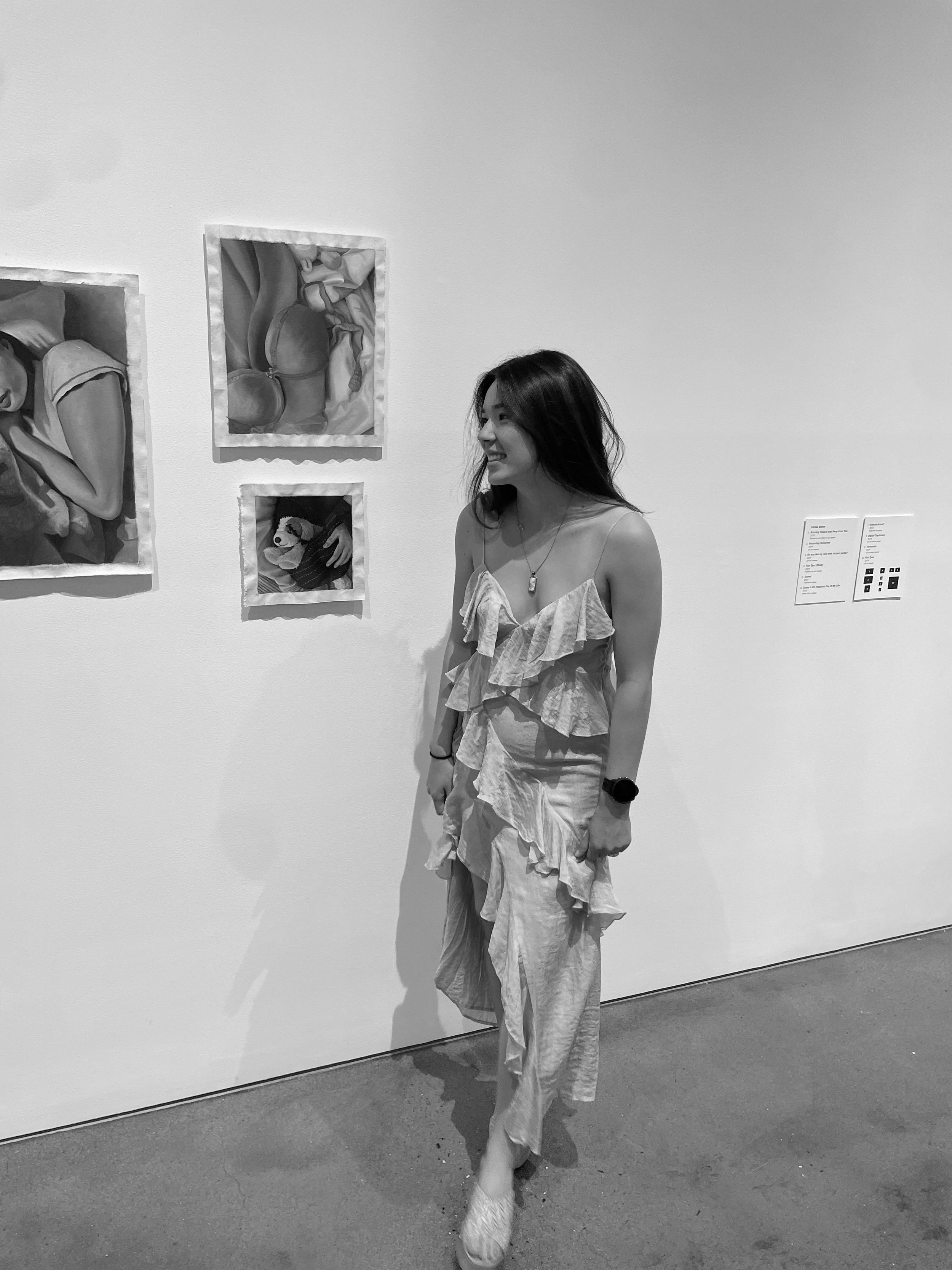 A young woman with long dark hair smiling and looking to her right, standing in an art gallery. She is wearing a flowy, ruffled dress and heels. On the gallery wall behind her, there are three framed paintings, one of a person sleeping, another of a still life with a  and objects, and a third of astuffed animal.