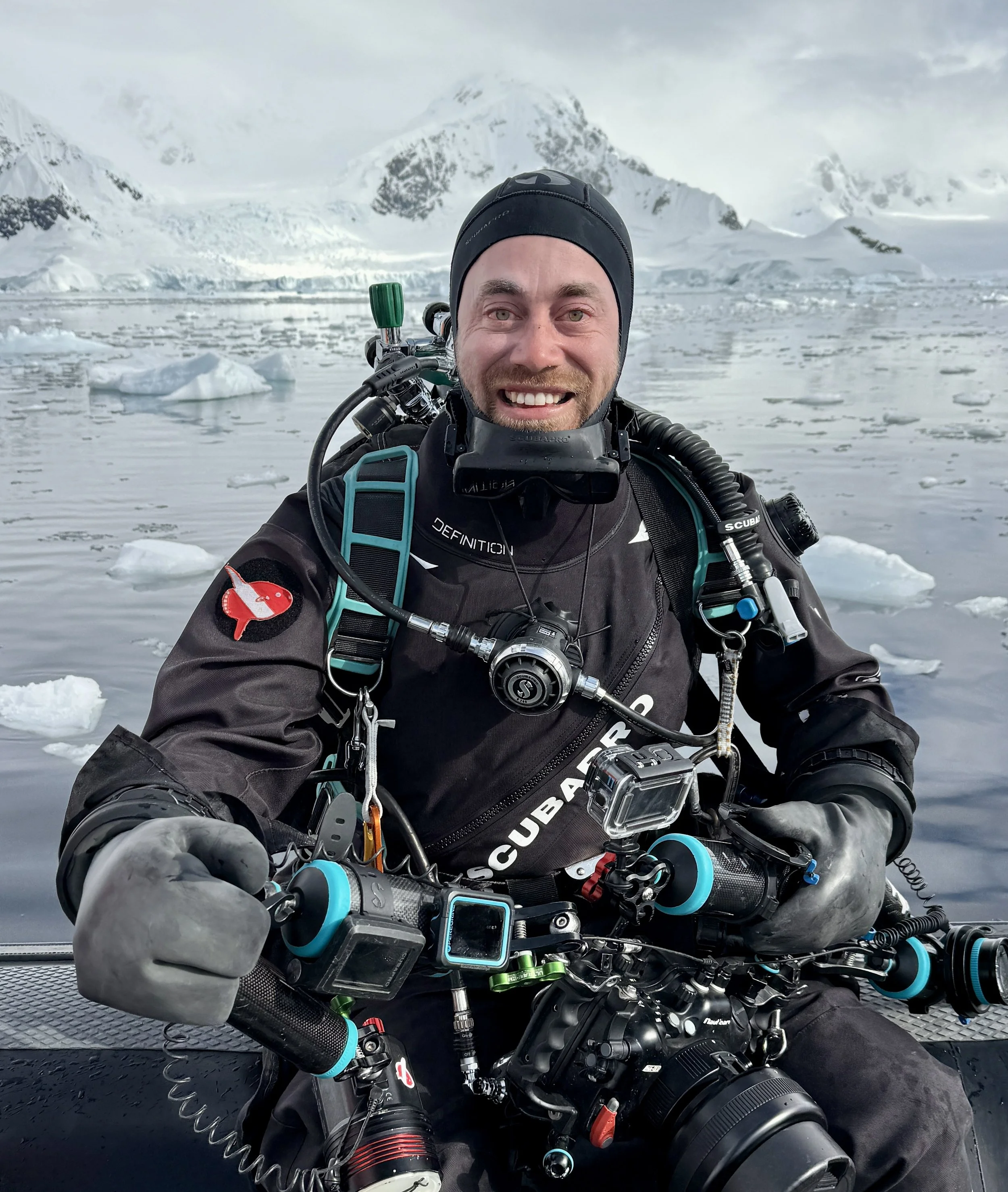 A man dressed in scuba diving gear holding a camera underwater in a frozen icy landscape with snow-covered mountains in the background.