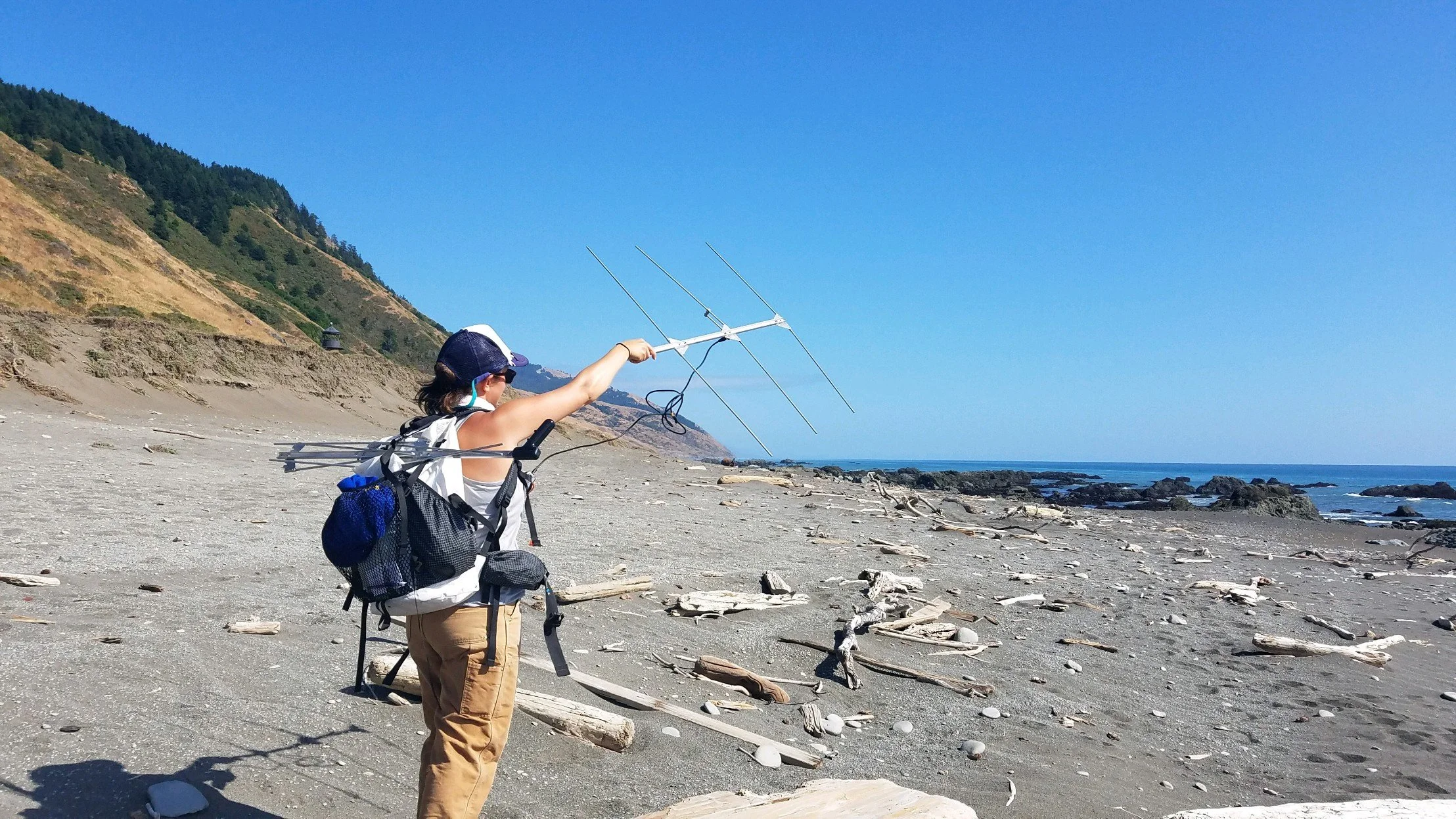 Person on a beach holding a large radio antenna, with a hillside and clear blue sky in the background.