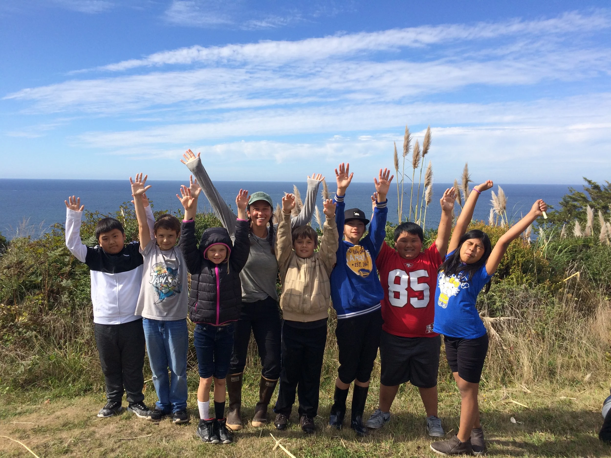 A group of children and a woman standing outdoors on a hillside, raising their hands in the air with the ocean and blue sky in the background.