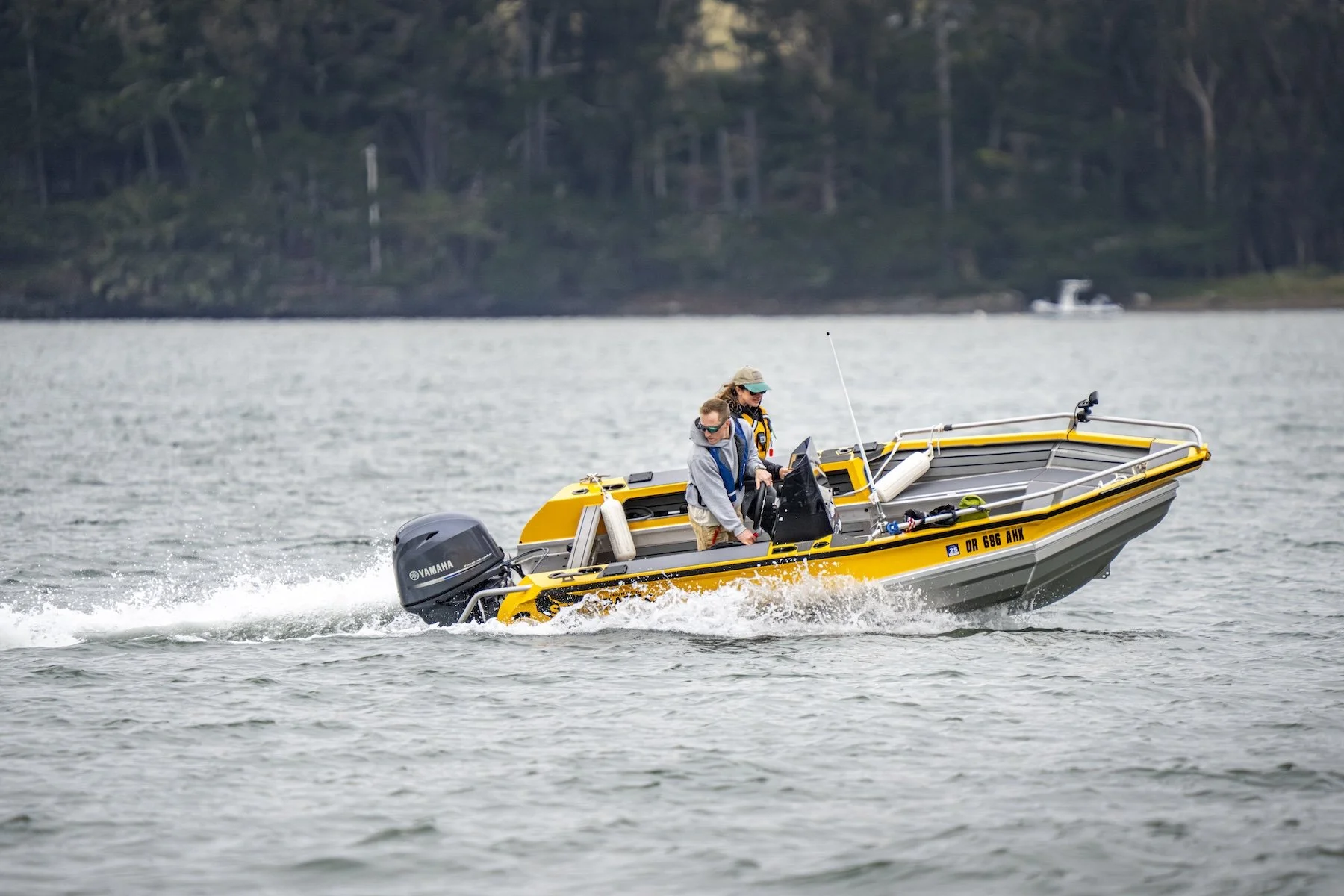A yellow motorboat with two people on board, navigating on a body of water, with trees and a boat in the background.