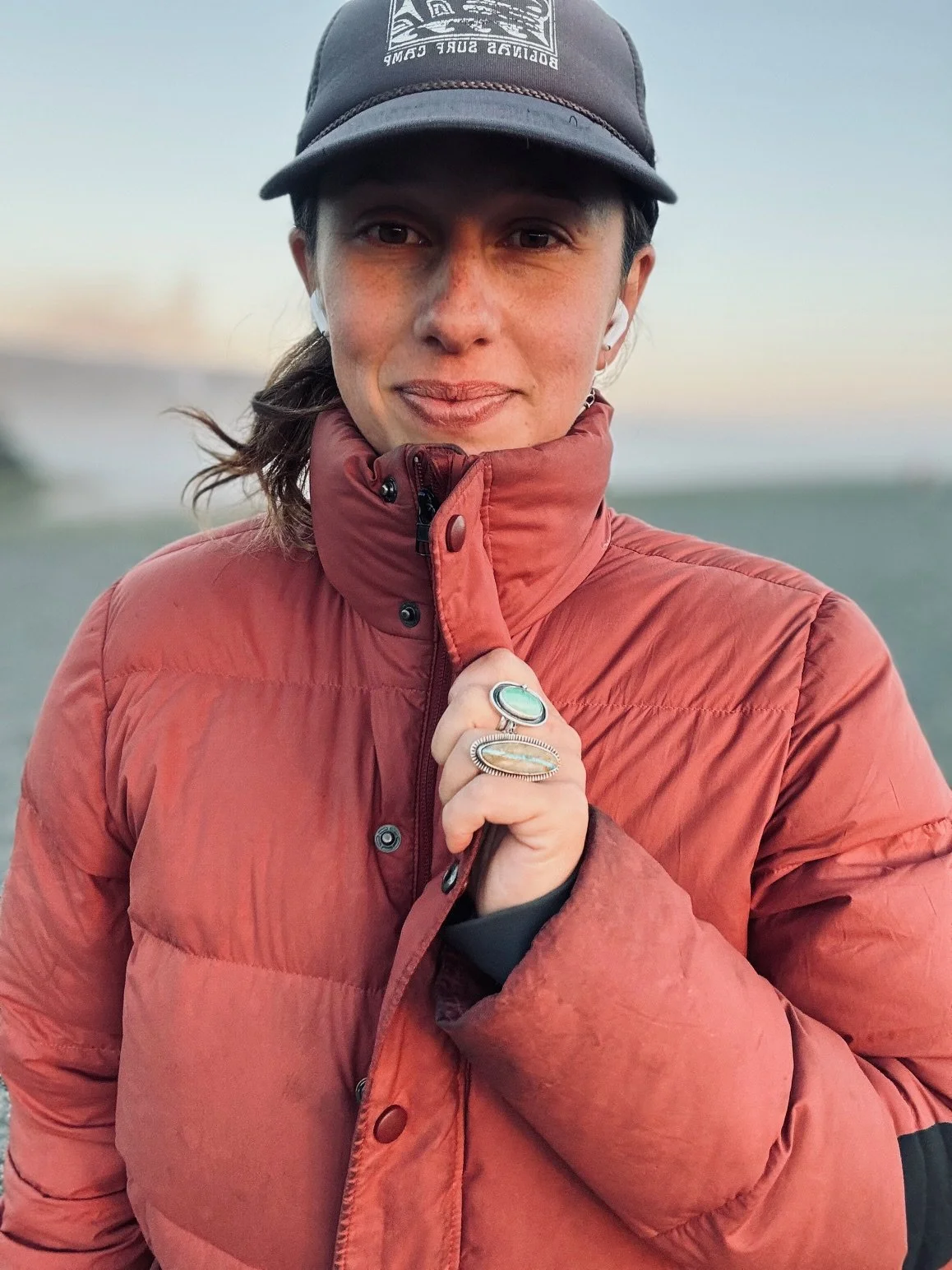 A woman outdoors in a red puffer jacket and dark cap, holding the collar of her jacket with her right hand, showing rings on her fingers, with a blurred natural background.