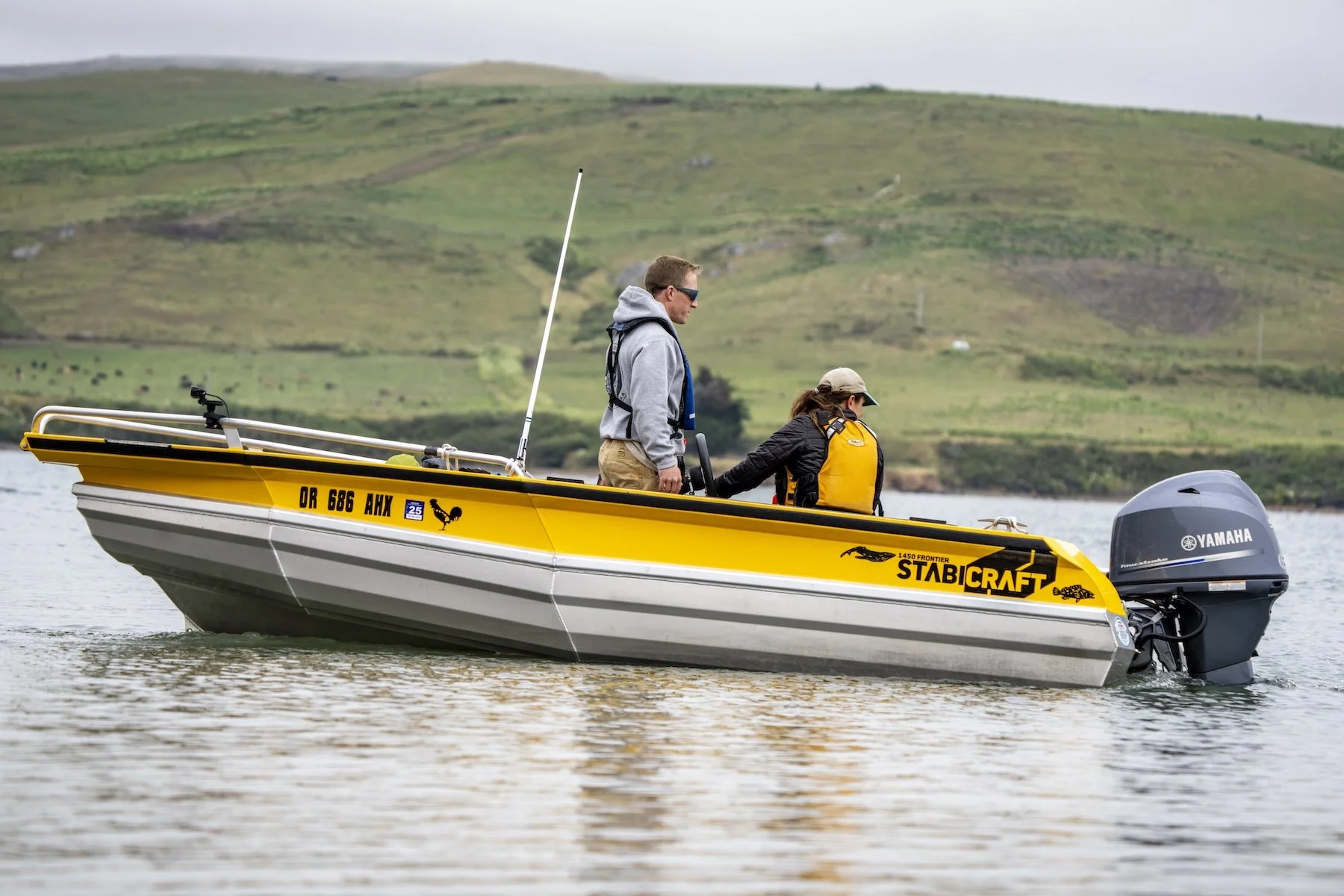Two people on a yellow and gray Stabicraft boat on calm water with green hills in the background. One person is sitting and the other is standing, both wearing life jackets. The boat has a Yamaha outboard motor.