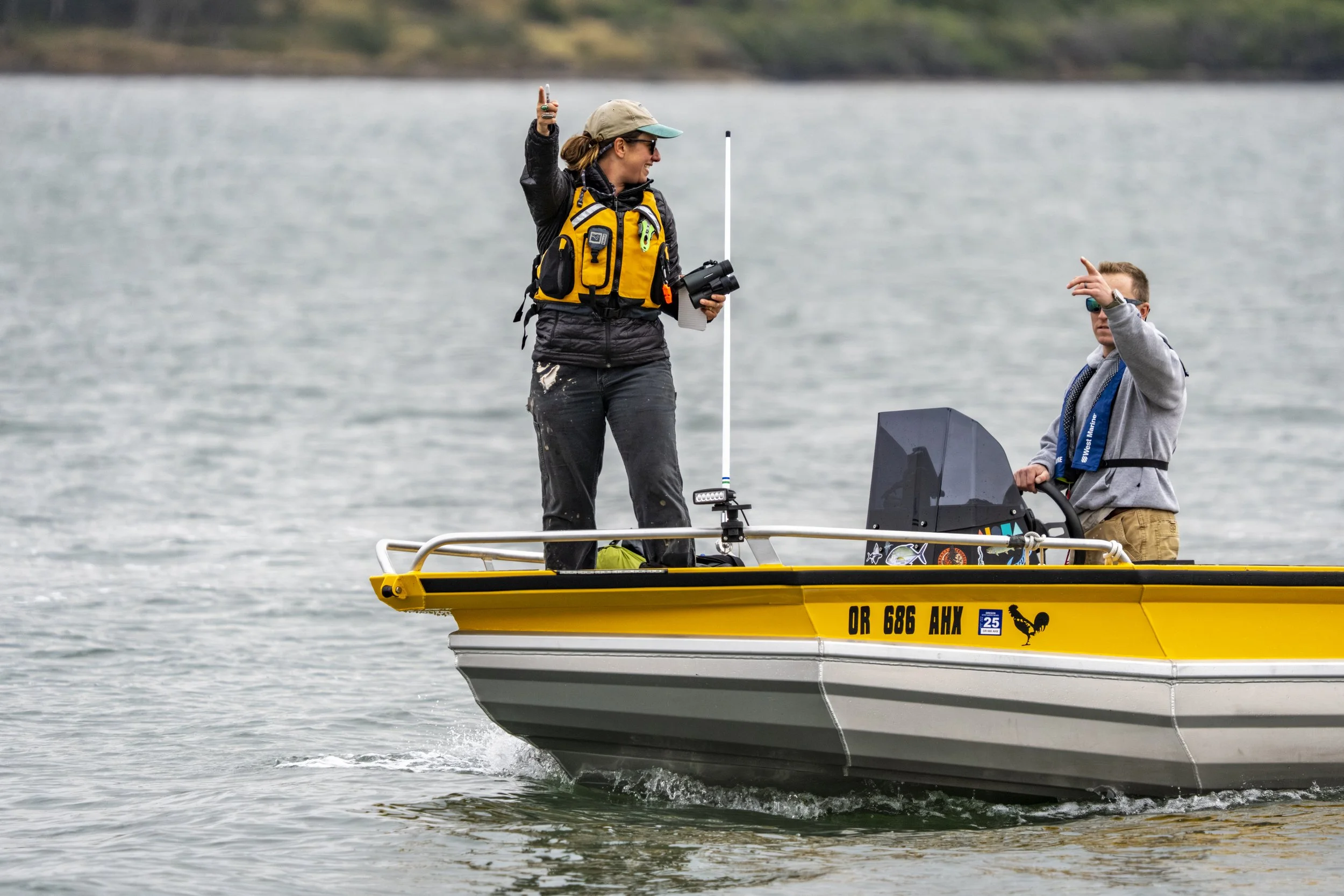 A woman wearing a yellow life vest, a beige cap, and sunglasses is on a yellow boat, holding a camera and a pole, with a young man in sunglasses and a gray hoodie sitting nearby near a body of water.