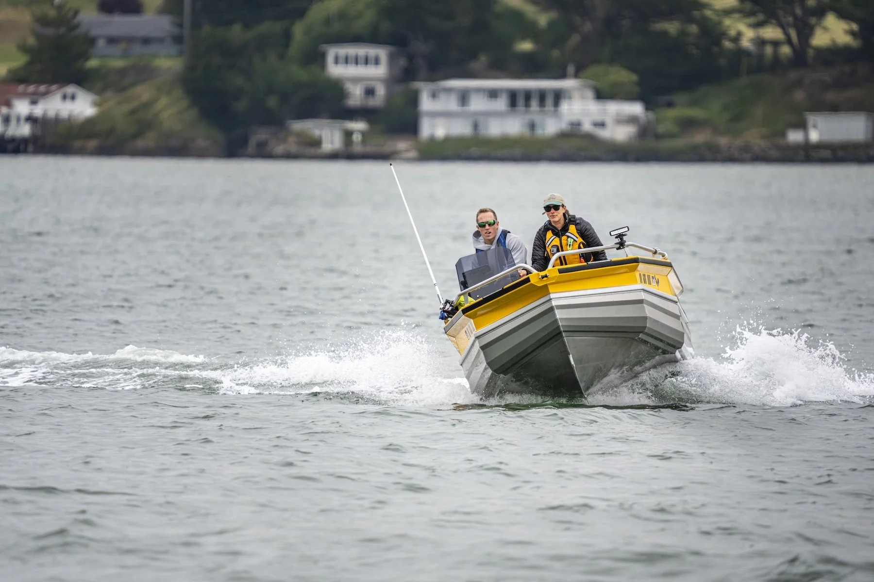 Two people in a yellow and gray motorboat riding on a lake, with houses visible on a hillside in the background.