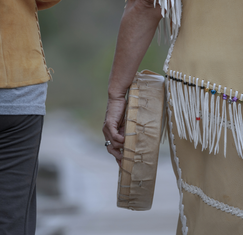 Close-up of a person's hand holding a drum, standing next to another person dressed in traditional Native American attire with fringe and beadwork.