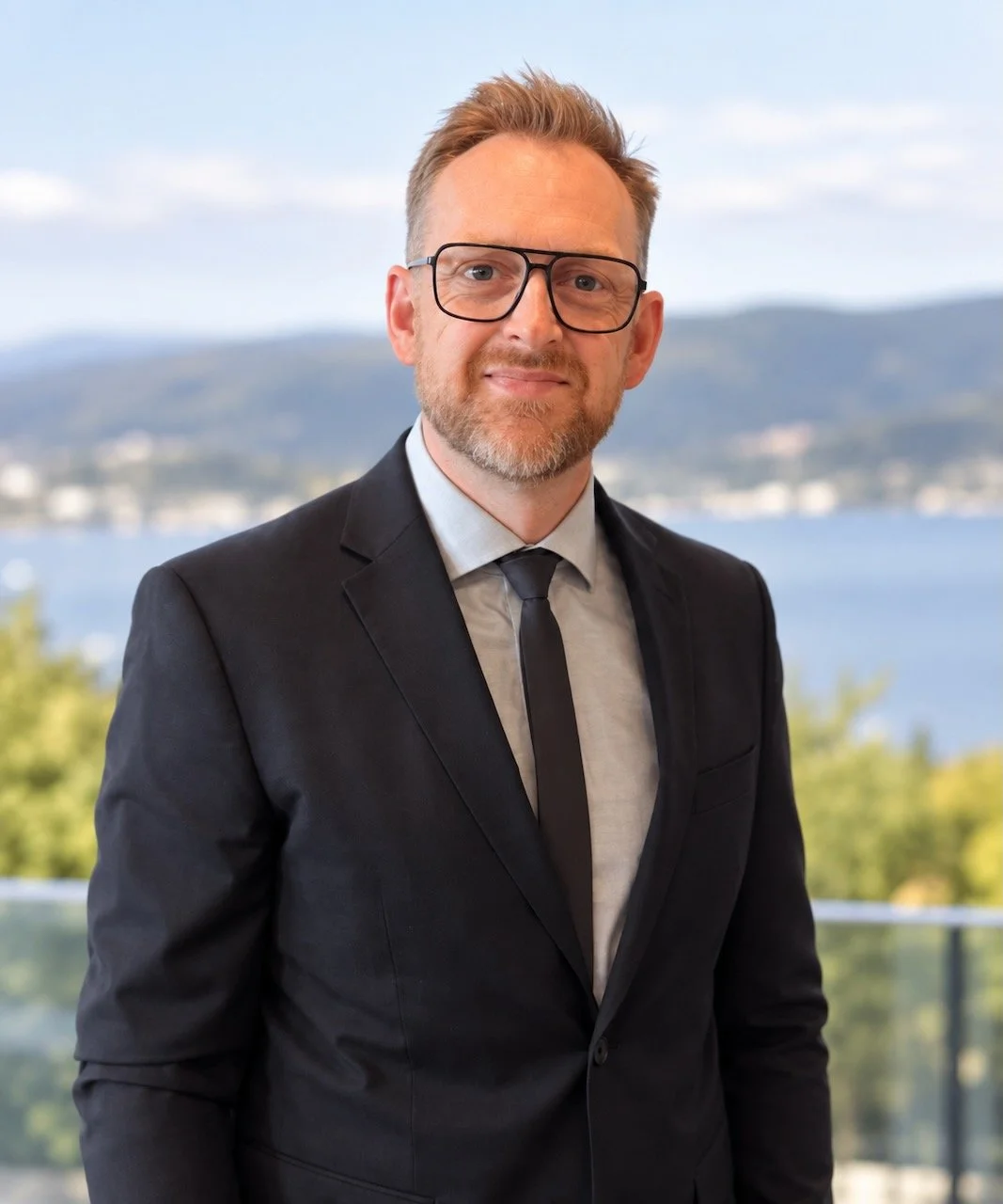 A man in a black suit, white shirt, and black tie standing outdoors with a scenic lake and mountains in the background.