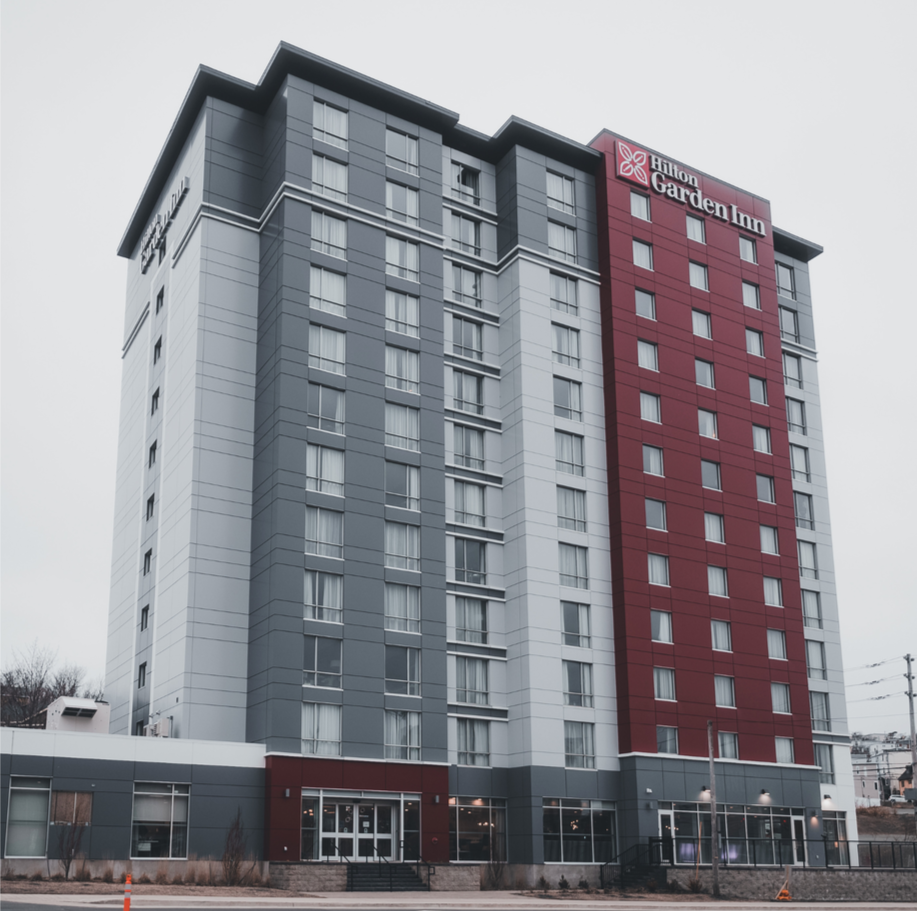 Exterior of a Hilton Garden Inn hotel, a tall modern building with gray and maroon panels, multiple windows, and a main entrance with glass doors.