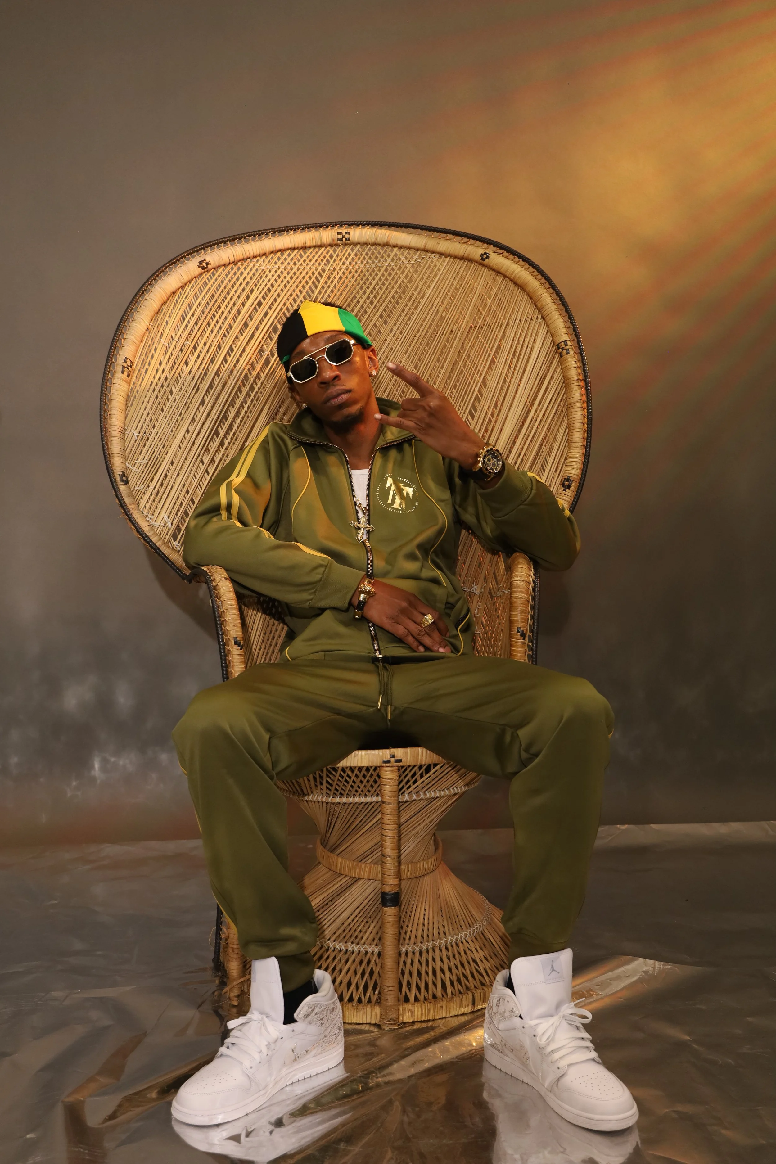 A young man sitting in a wicker chair against a plain background, wearing sunglasses, a green tracksuit, a colorful cap, white sneakers, and accessories, making a hand gesture with his right hand.
