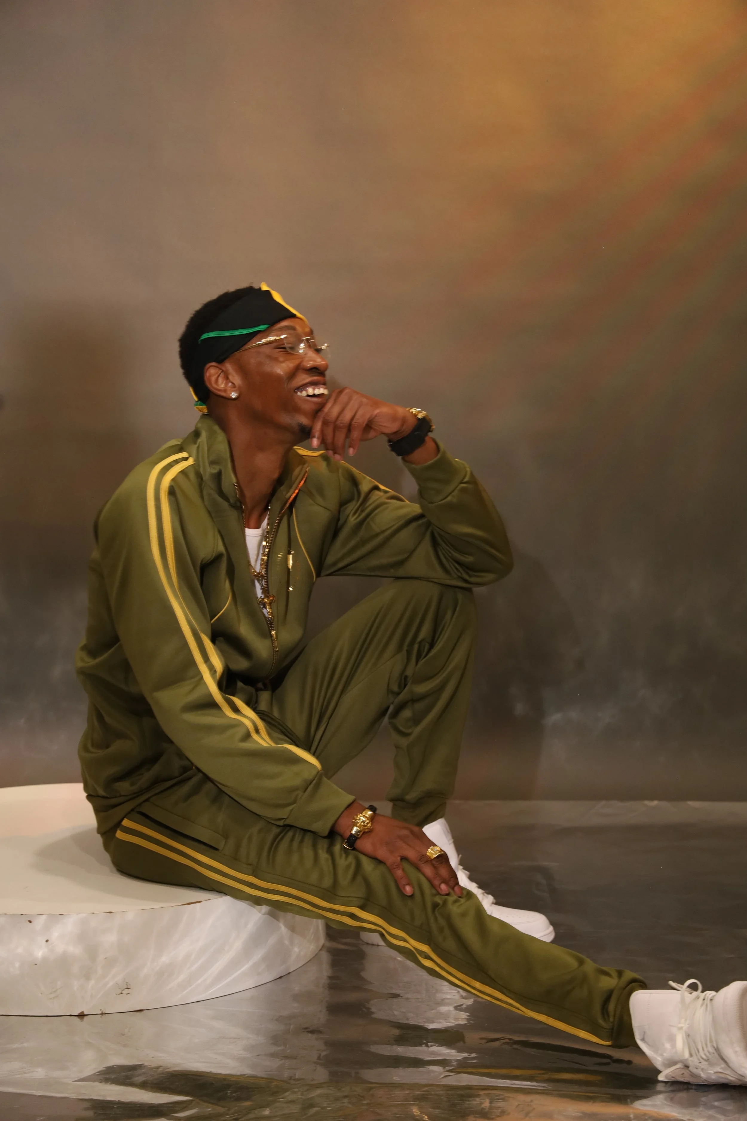 A man wearing a green tracksuit with yellow stripes, white sneakers, sunglasses, and a black headscarf with yellow accents sits on a marble surface against a gray wall, smiling and resting his arm on his knee.