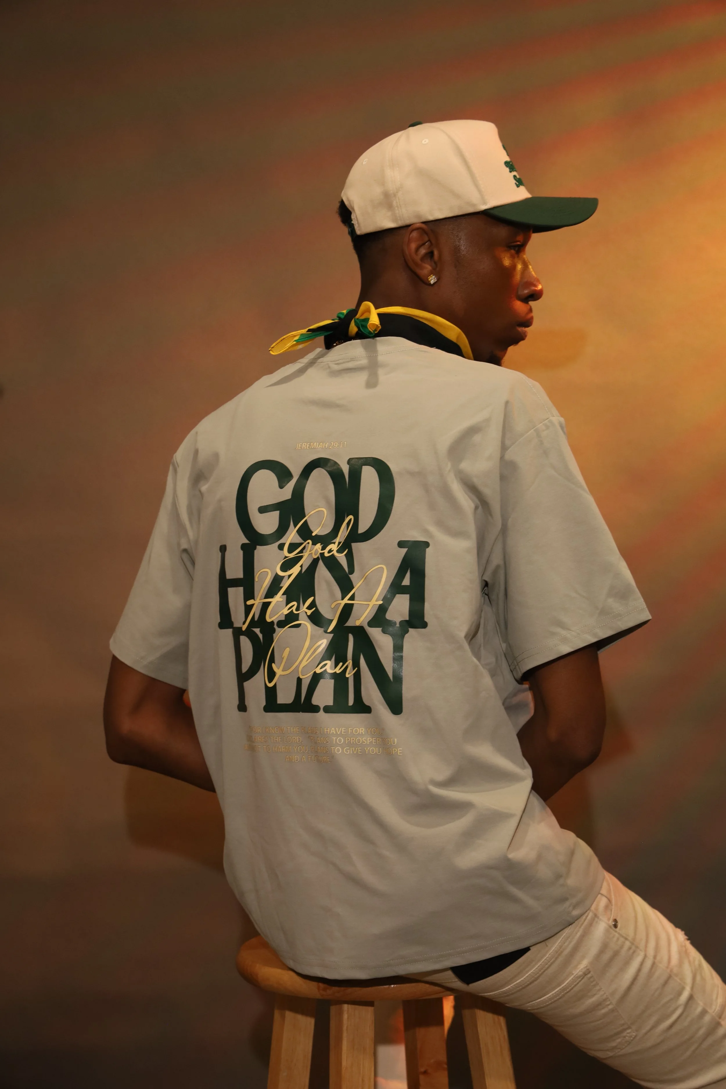 Person sitting on a wooden stool against a plain colored background, wearing a white baseball cap, earrings, a t-shirt with large text, and a yellow, black, and green neck scarf.