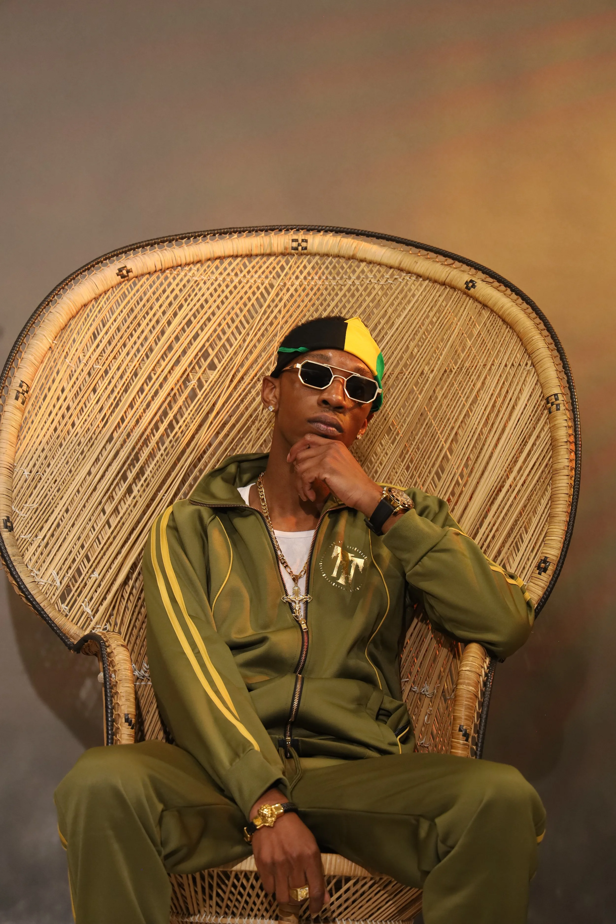 Person sitting on a large wicker chair, wearing sunglasses and a green tracksuit with yellow stripes, accessorized with jewelry, posed thoughtfully against a plain background.