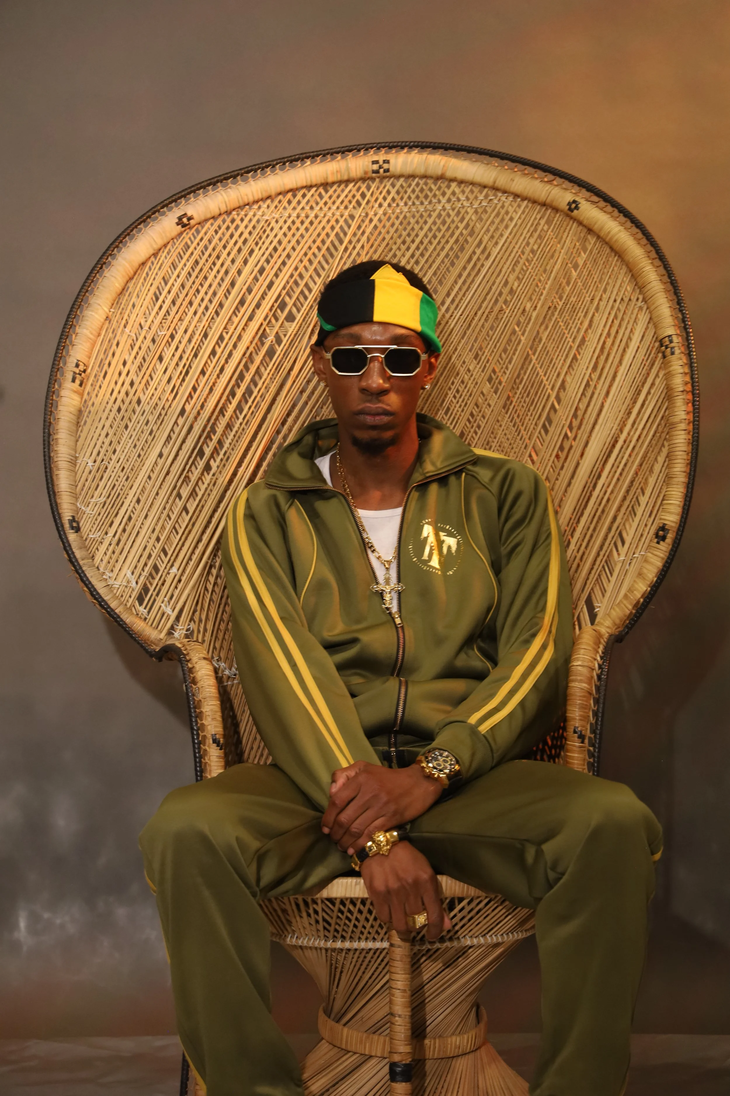 A man sitting in a large wicker peacock chair, wearing an olive green tracksuit with yellow accents, a multicolored headscarf, sunglasses, and gold jewelry. The background is a plain wall.
