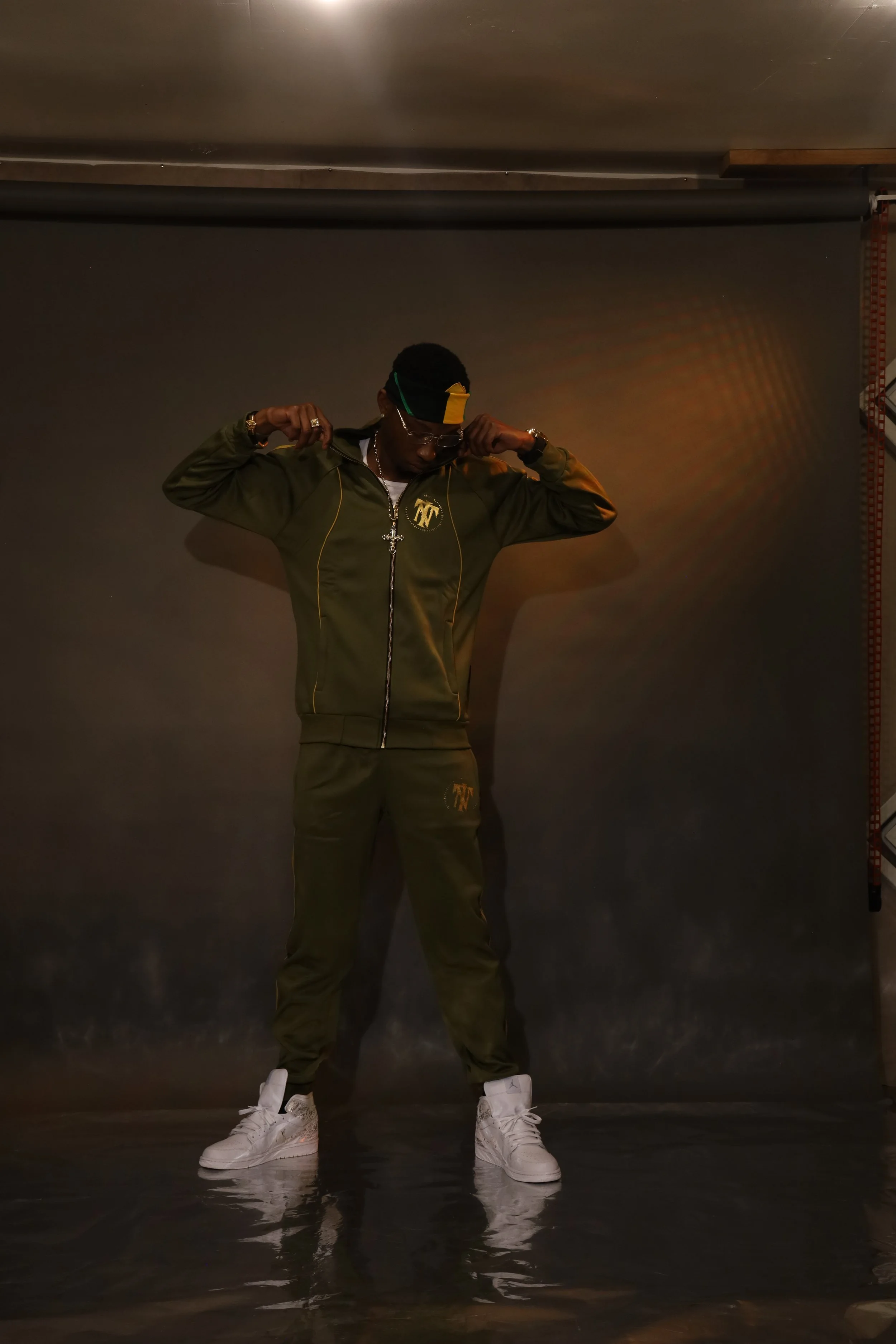 A man dressed in a green tracksuit and white sneakers standing against a plain dark background, adjusting his jacket.