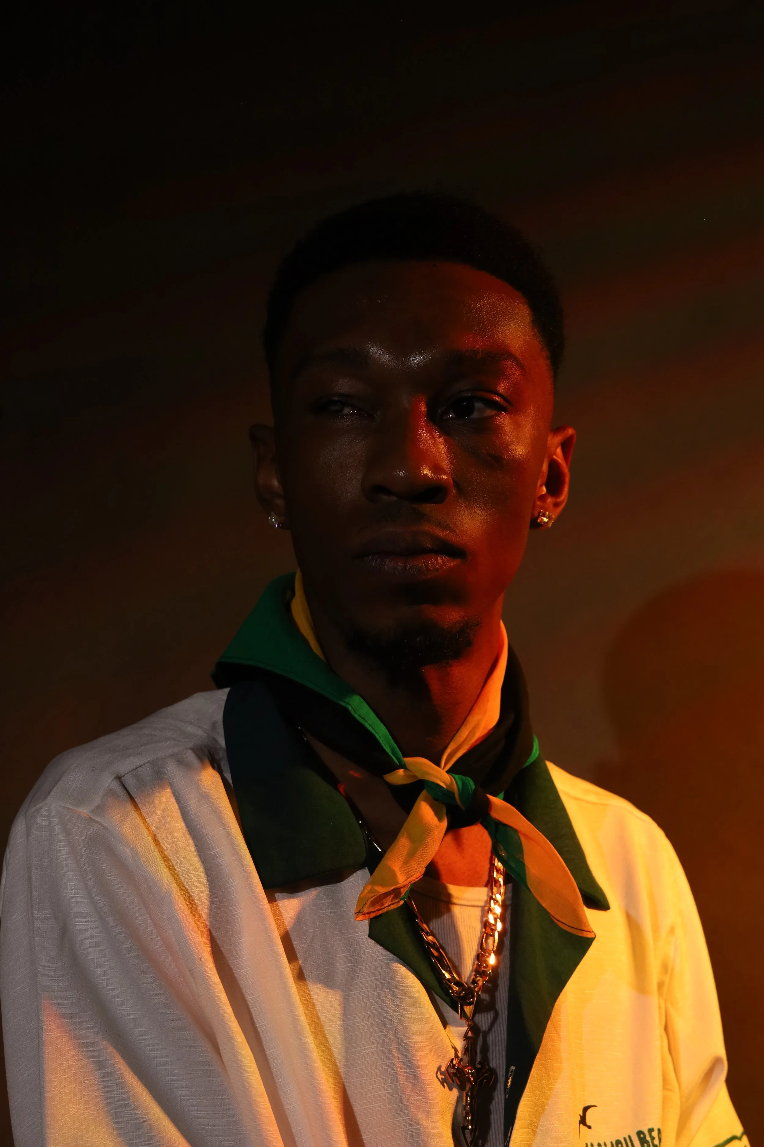 A young man with dark skin and short black hair, wearing earrings, a chain necklace, and a tan scout neckerchief with green accents, stands outdoors at night with a dark sky overhead.