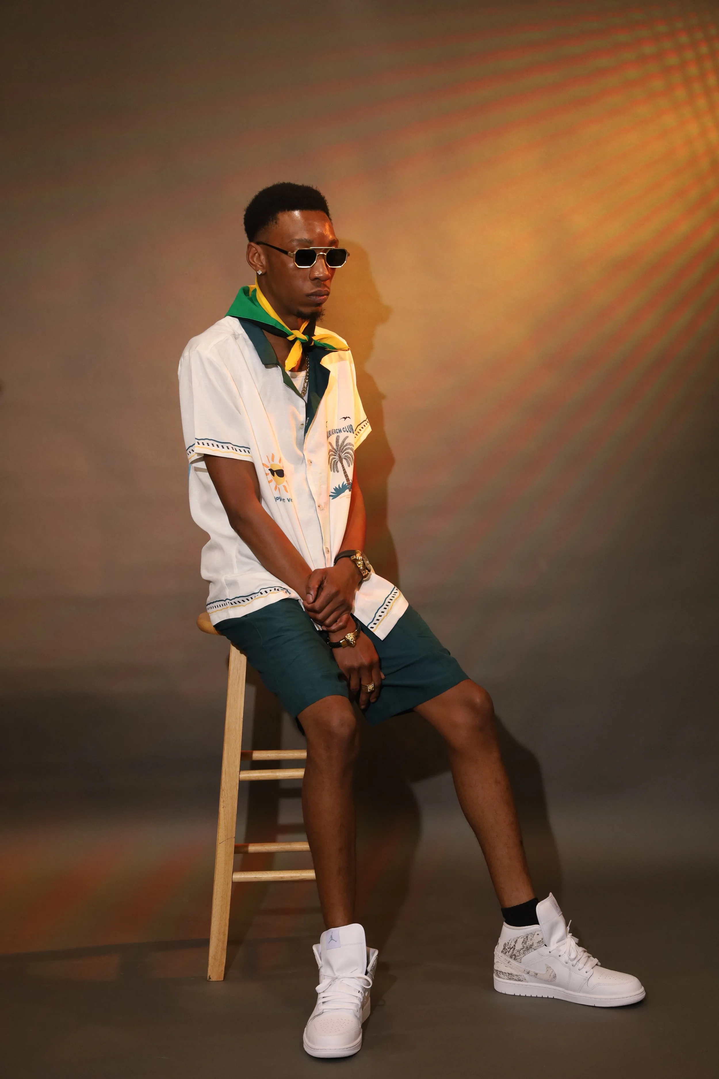 A young man sitting on a wooden stool against a plain wall. He is wearing sunglasses, a colorful tropical shirt, dark shorts, white sneakers, and has a green, yellow, and black bandana around his neck. He also has jewelry, including rings, earrings, a watch, and a chain. A shadow of his profile is cast on the wall behind him.
