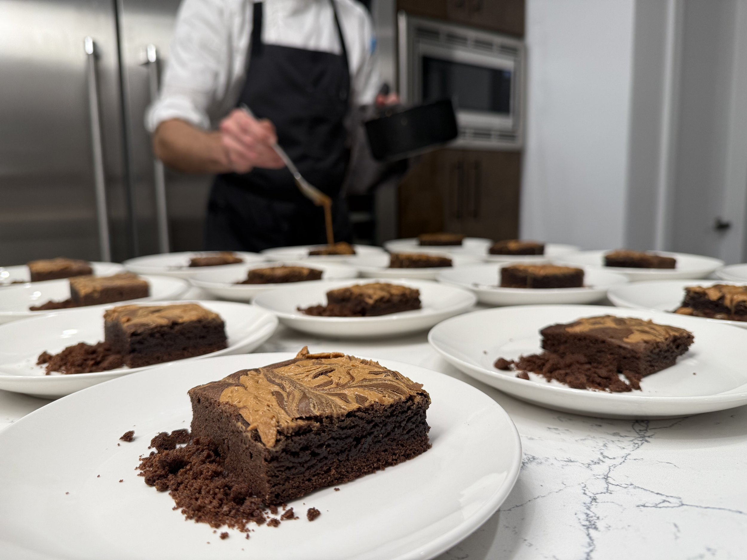 Close-up of a chocolate dessert with caramel topping on a white plate, with more similar desserts on plates in the background, in a kitchen setting.
