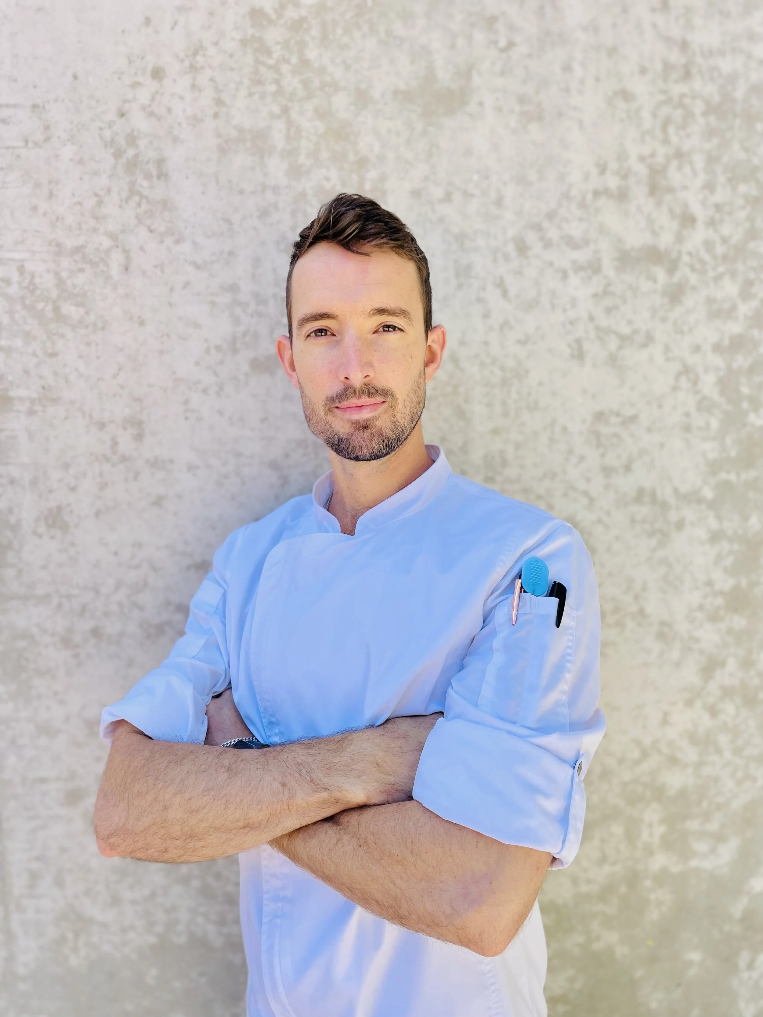 A male healthcare professional wearing a light blue medical uniform with pens in the pocket, standing outdoors against a textured beige wall with arms crossed.