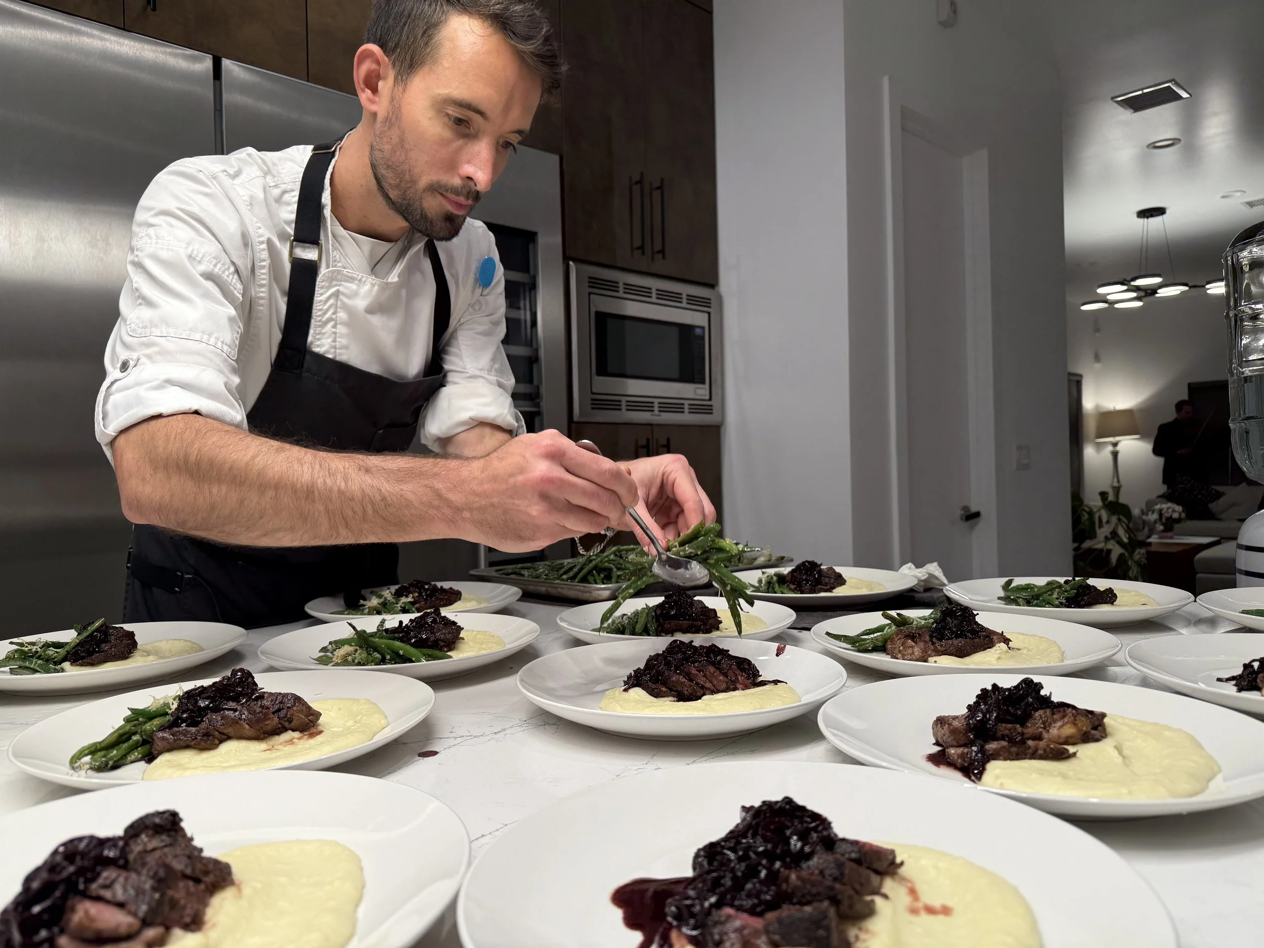 A chef in a white shirt and black apron preparing plates of food in a modern kitchen. The plates contain mashed potatoes, grilled meat topped with dark sauce, and green beans.