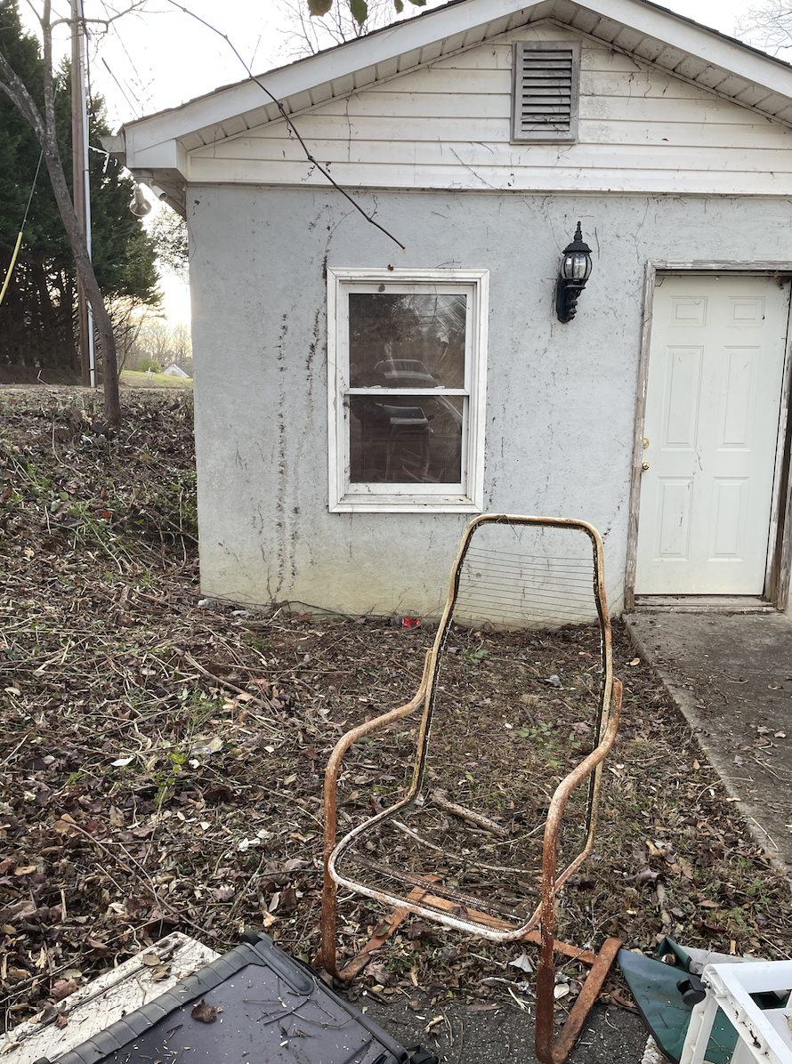 A rusty outdoor chair with a mesh seat and backrest in front of a house wall with a window and a lamp, surrounded by dry leaves and debris.