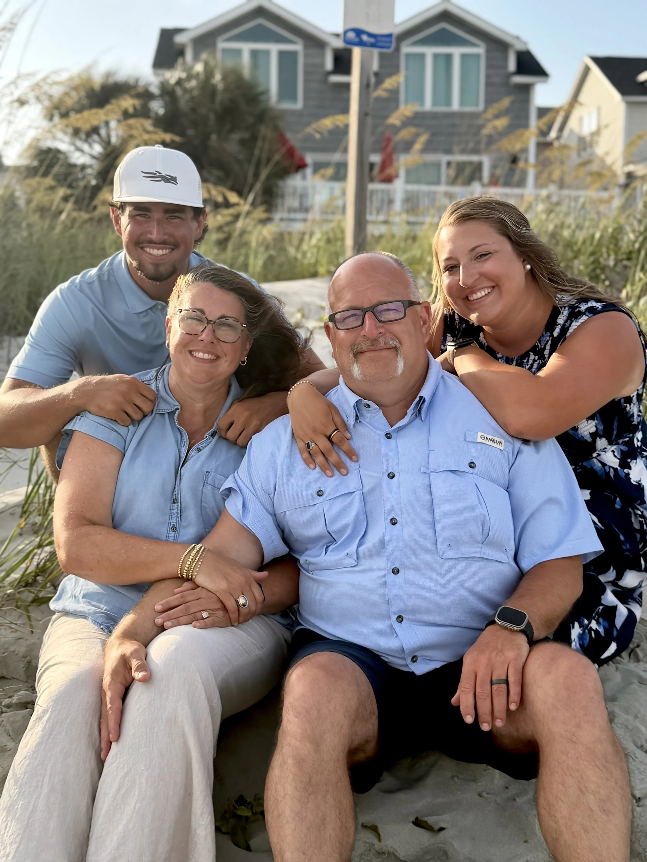 A group of five people sitting on sand dunes near beachfront houses, smiling at the camera. They are dressed casually, with three women and two men, one wearing glasses and the other in a cap.