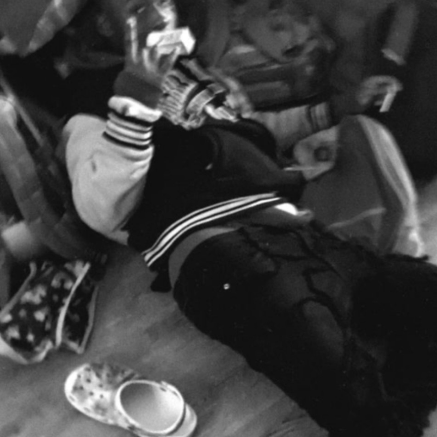 Child sitting on the floor, wearing striped socks and Crocs with a pattern, next to a pair of slip-on shoes, surrounded by various objects.