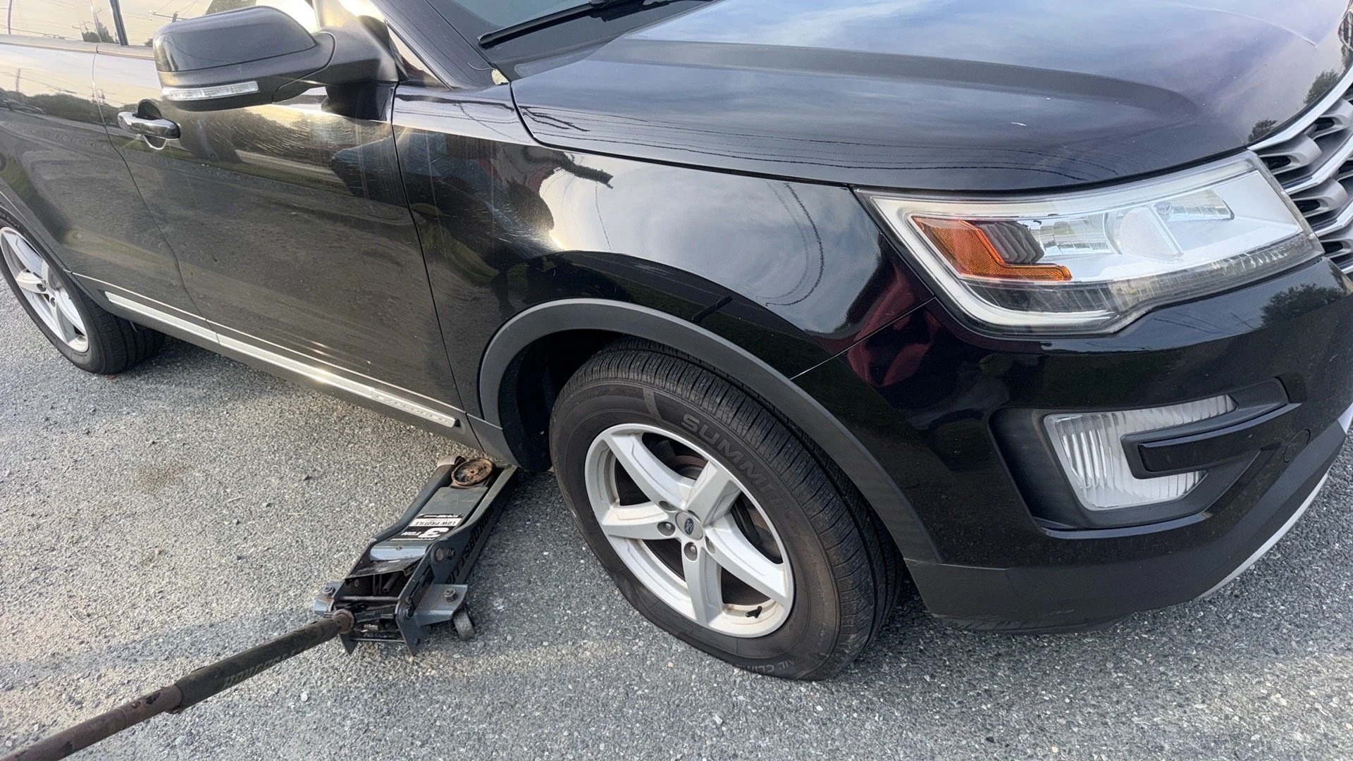 Black SUV vehicle parked on a gravel surface with a hydraulic floor jack underneath the front passenger side. An automotive tool is positioned near the front tire.