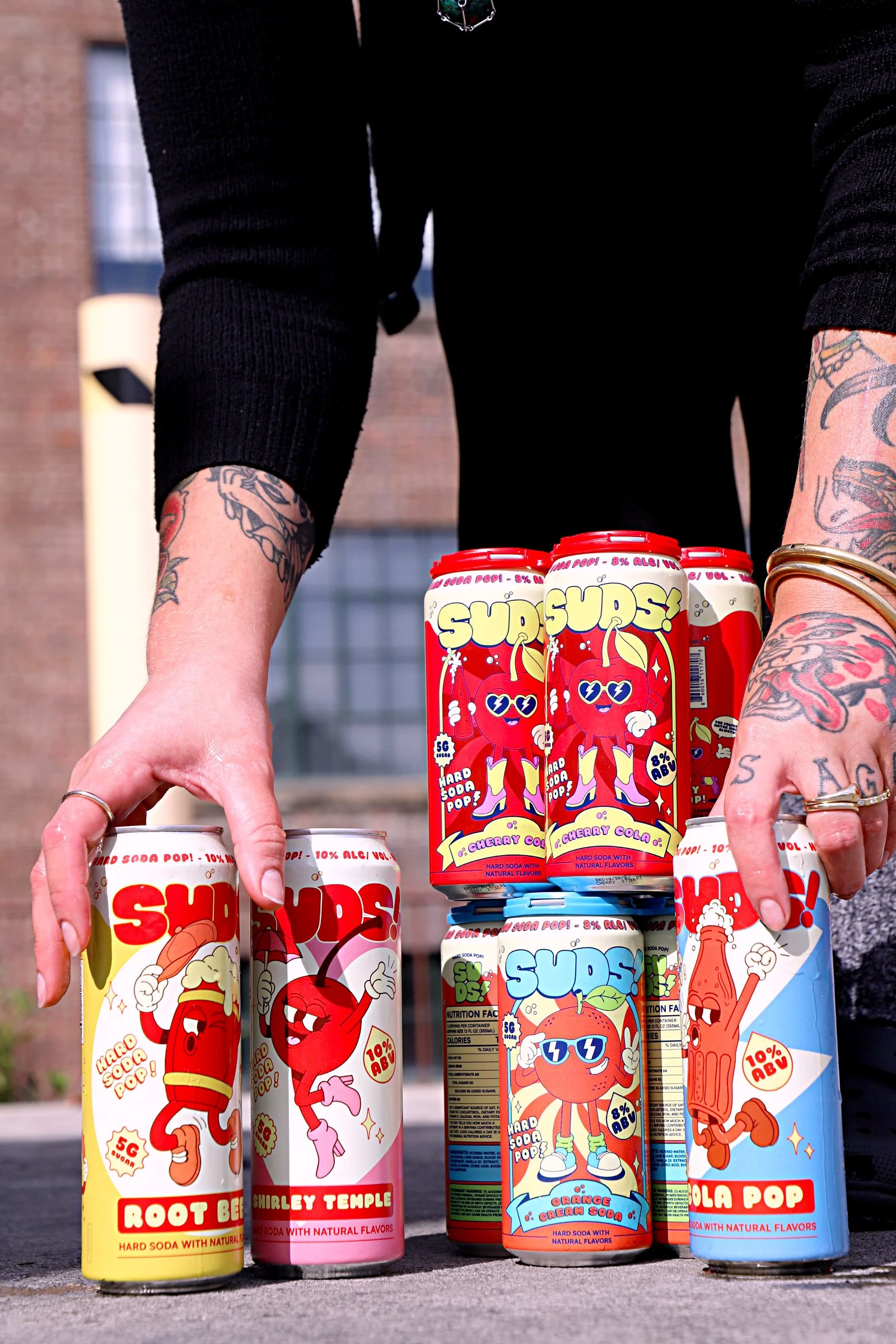 Several colorful cans of soda are arranged on a the ground outdoors, with a person reaching out to touch one of them. The cans feature cartoon characters and vibrant designs, with flavors such as cherry cola, orange cream, root beer, and cola pop.