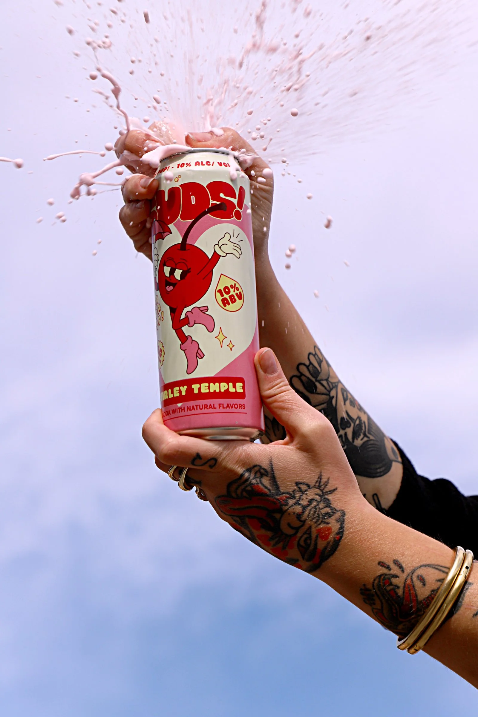 A person with tattoos on their arm opening a can of Shirley Temple Hard Soda Pop, causing pink liquid to splash out against a plain sky background.