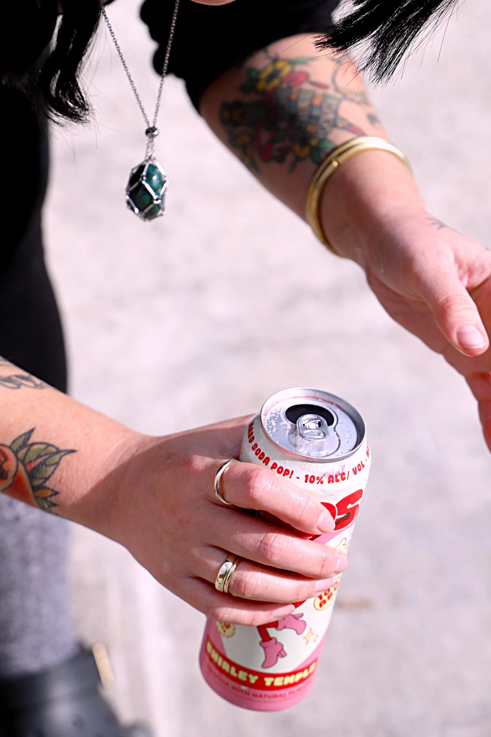 A person's hands holding an open can of soda with a pink and white label, with tattoos on their arms and wearing gold jewelry and a necklace with a large pendant.