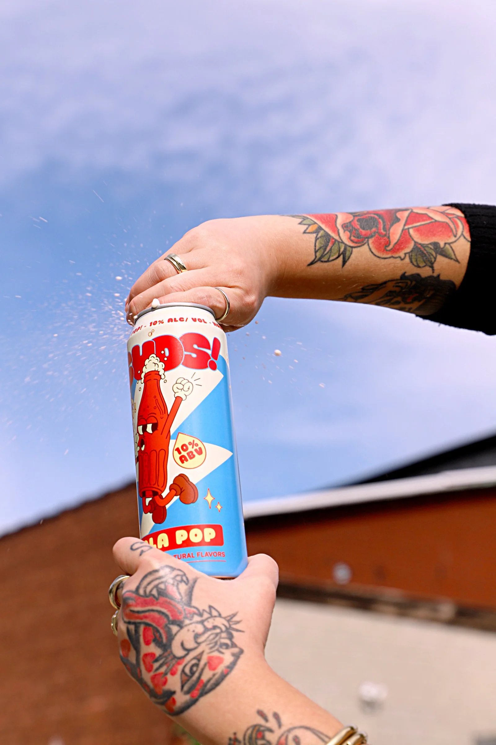Person with tattooed arm holding a can of La Pop soda, with the soda spraying out against a clear sky background.
