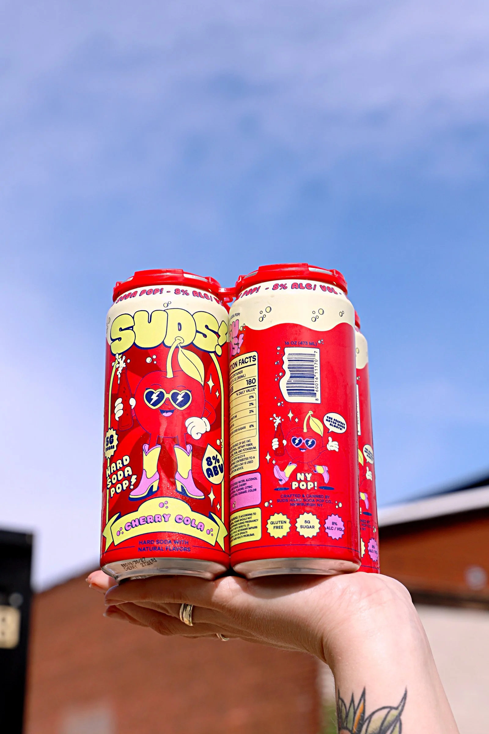 Two cans of cherry cola soda with bright red packaging and cartoon cherry character on a person's hand against a blue sky background.