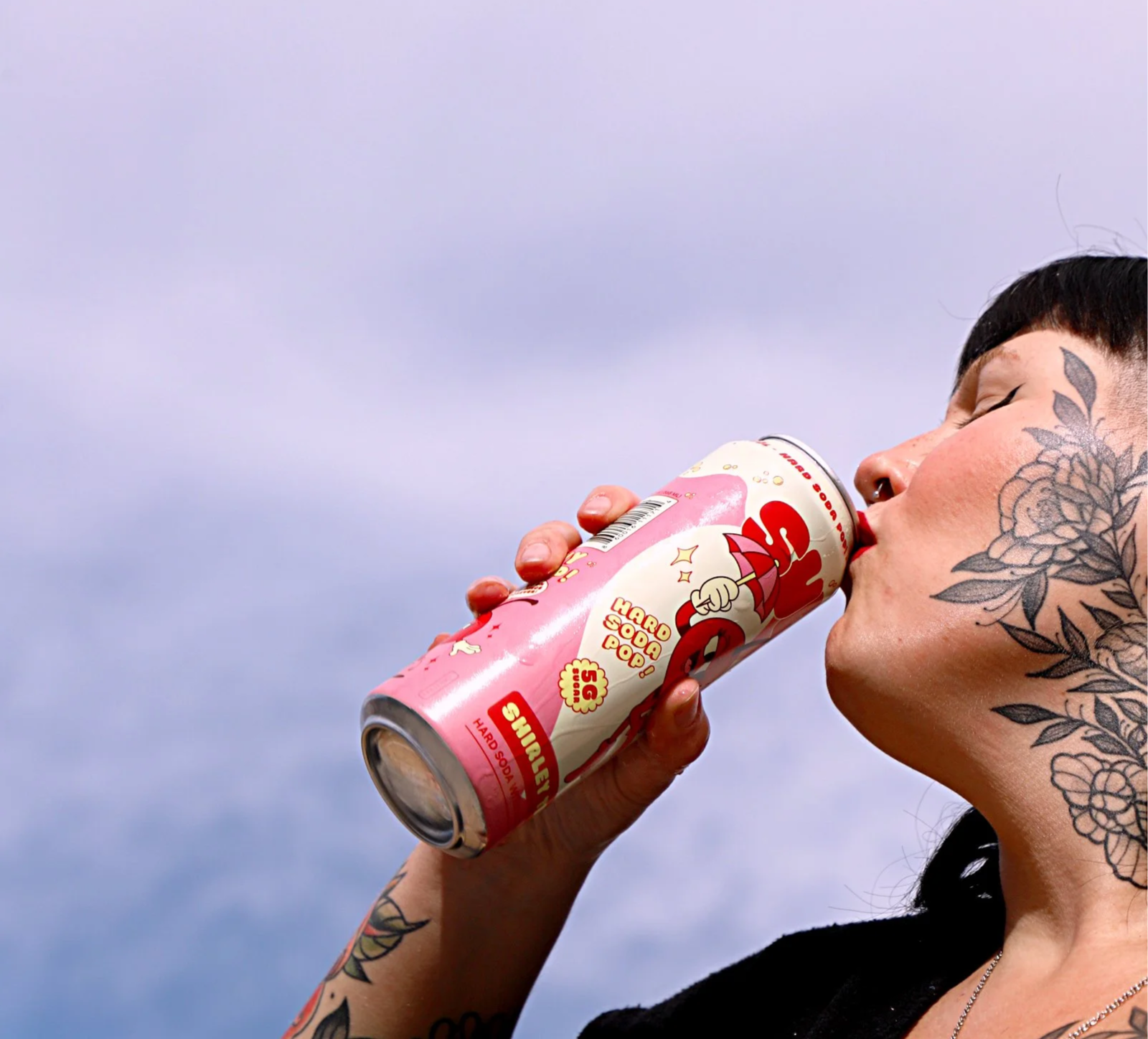 A woman with tattoos on her face and arm drinks from a can of Shirley Temple Hard Soda Pop against a cloudy sky background.
