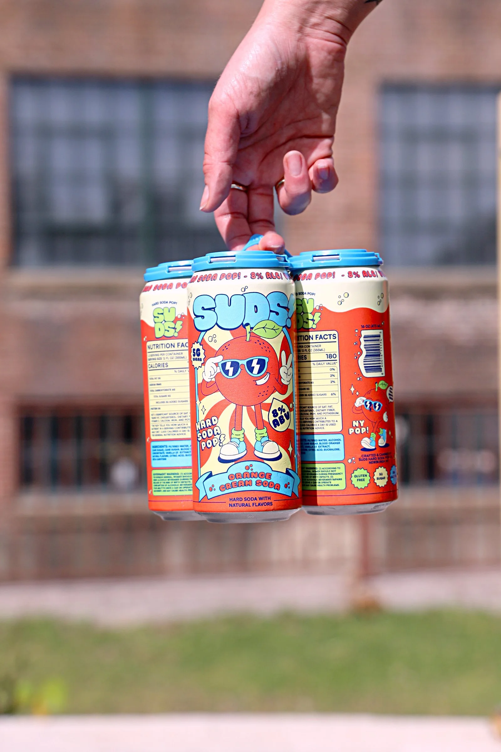 Hand holding three cans of SUDS! orange cream soda with playful cartoon character on the label, set against a blurred building background.