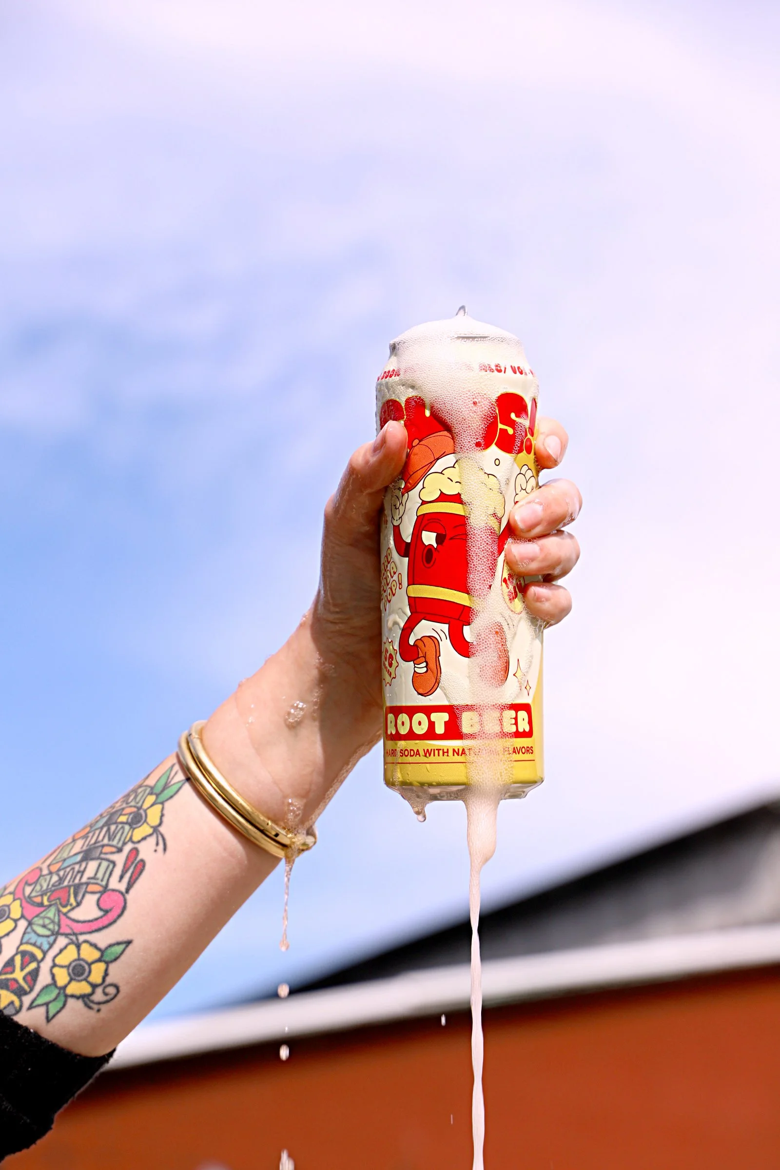 A hand holding a can of root beer soda with foam overflowing from the top, against a sky background.