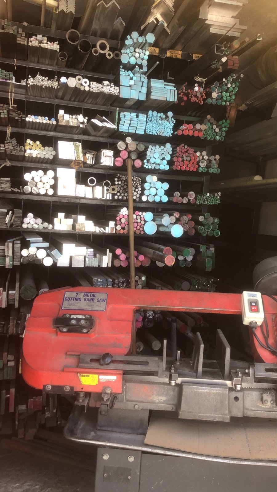 Estante con barras de metal de diferentes tamaños y colores almacenadas en un taller, con una sierra de banda metálica de 7 pulgadas al frente.