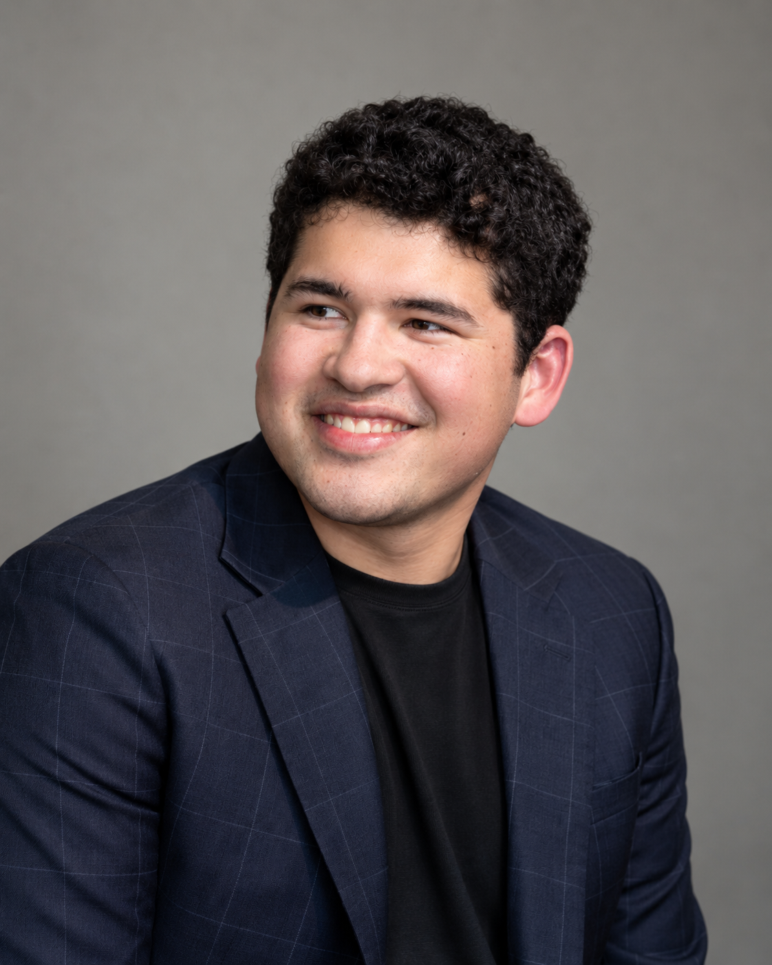 Young man with dark, curly hair, smiling, wearing a dark blazer over a black shirt, against a neutral gray background.