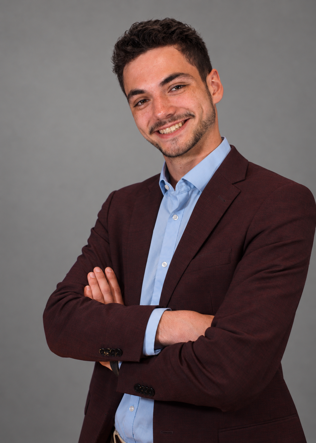 A young man with dark, curly hair and a beard, dressed in a maroon blazer and light blue shirt, smiling with arms crossed against a gray background.