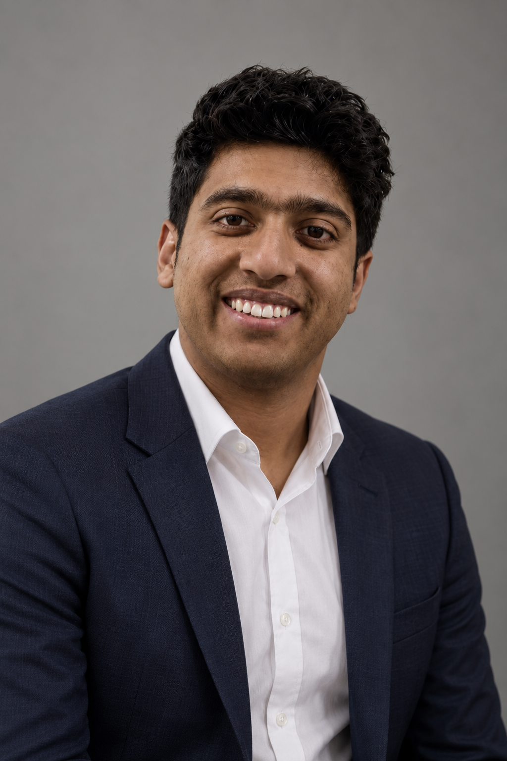 Professional headshot of a smiling man with dark hair in a suit and white shirt against a plain gray background.