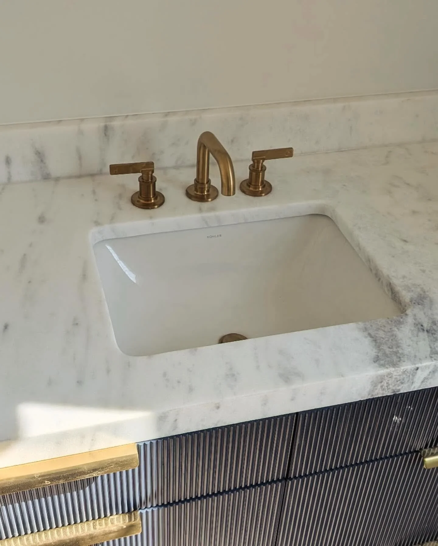 A bathroom sink with a white marble countertop, a central gold faucet, and two gold handles for hot and cold water.