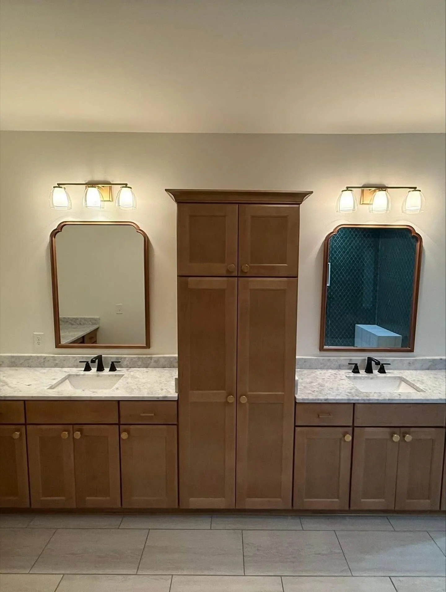 Double bathroom vanity with two mirrors, two sets of three-light fixtures, and black faucets, with wooden cabinets and drawers, and a tall wooden storage cabinet in the center.