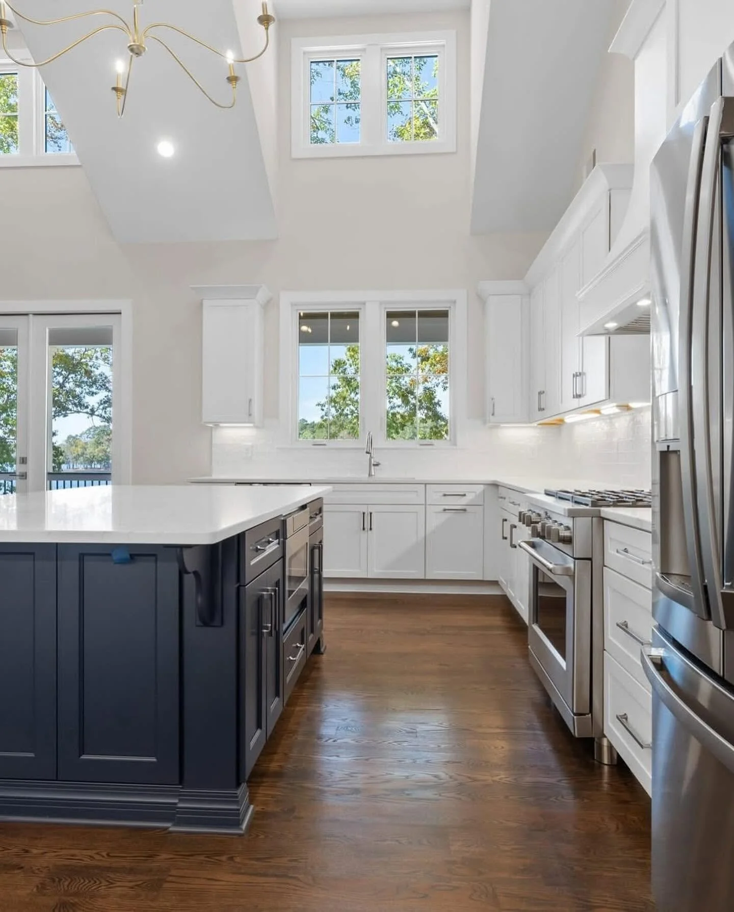 Bright kitchen with white cabinets, a kitchen island with gray base, stainless steel appliances, hardwood floor, and large windows letting in natural light.