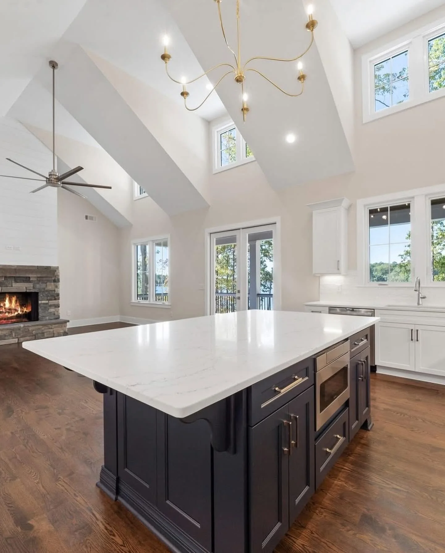 Modern kitchen with white cabinets, a large white marble island, dark cabinet base, and hardwood flooring. Features a stone fireplace, multiple windows, and a high vaulted ceiling with a gold chandelier and ceiling fan.