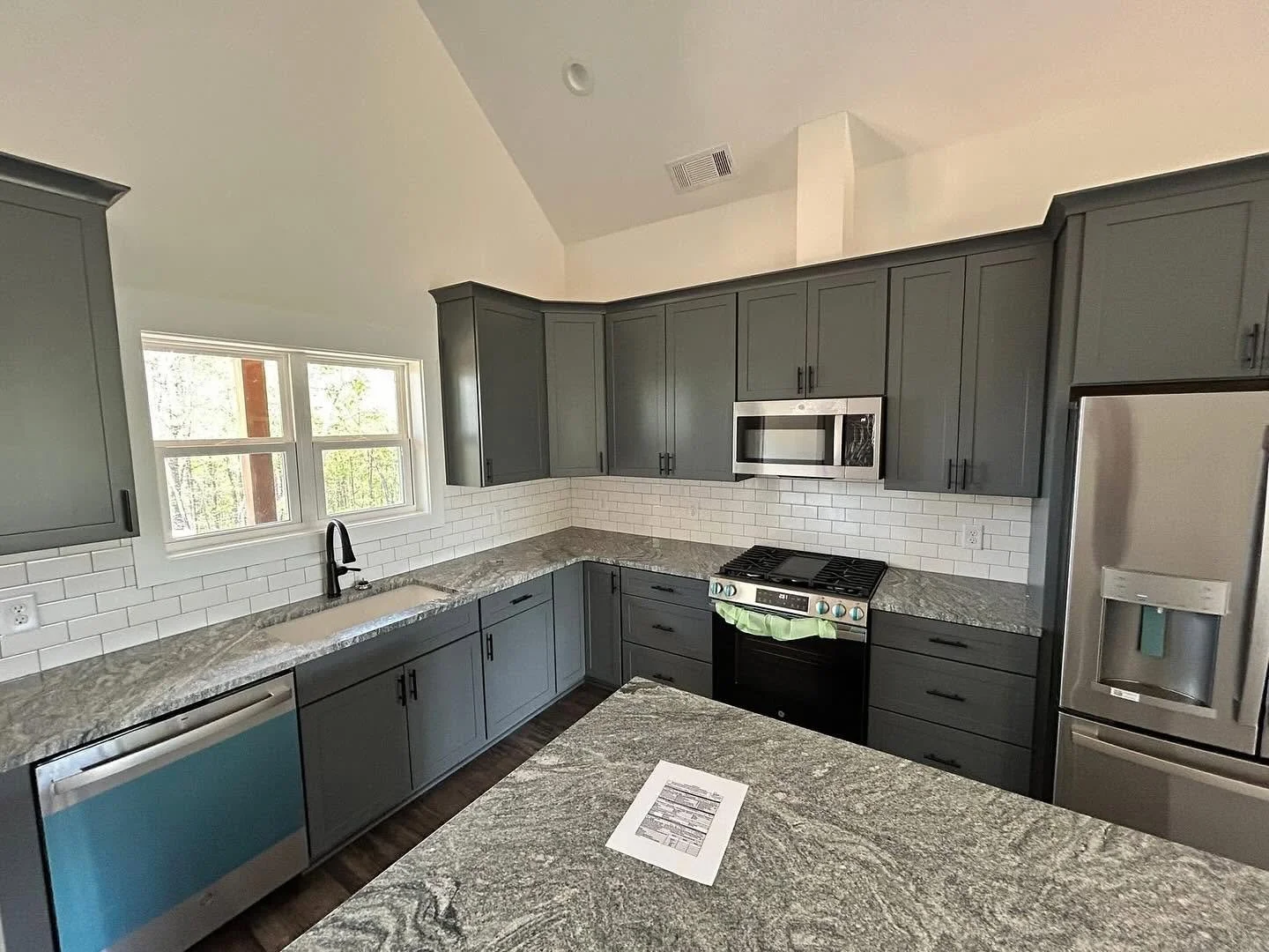 Modern kitchen with gray cabinets, granite countertops, white subway tile backsplash, stainless steel appliances including a microwave and refrigerator, and a window above the sink.
